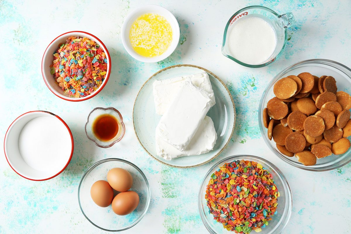 Ingredients for Taste of Home's Fruity Pebbles Cheesecake laid out in small bowls on a blue and green surface.