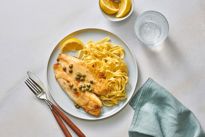 Tilapia fillet served with pasta and lemon butter sauce on a serving plate with a lemon wedge on the side