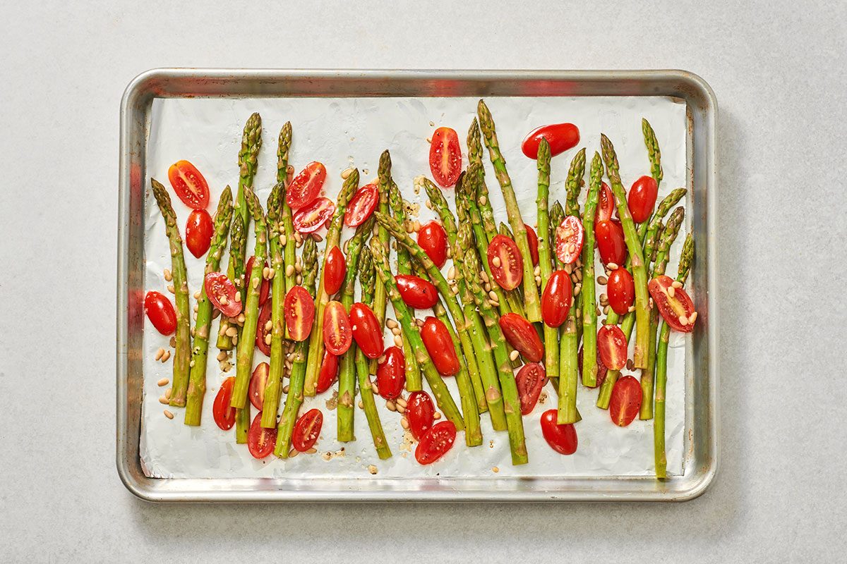 Asparagus tomatoes and pine nuts tossed with olive oil salt and pepper sauce on a rimmed baking pan.