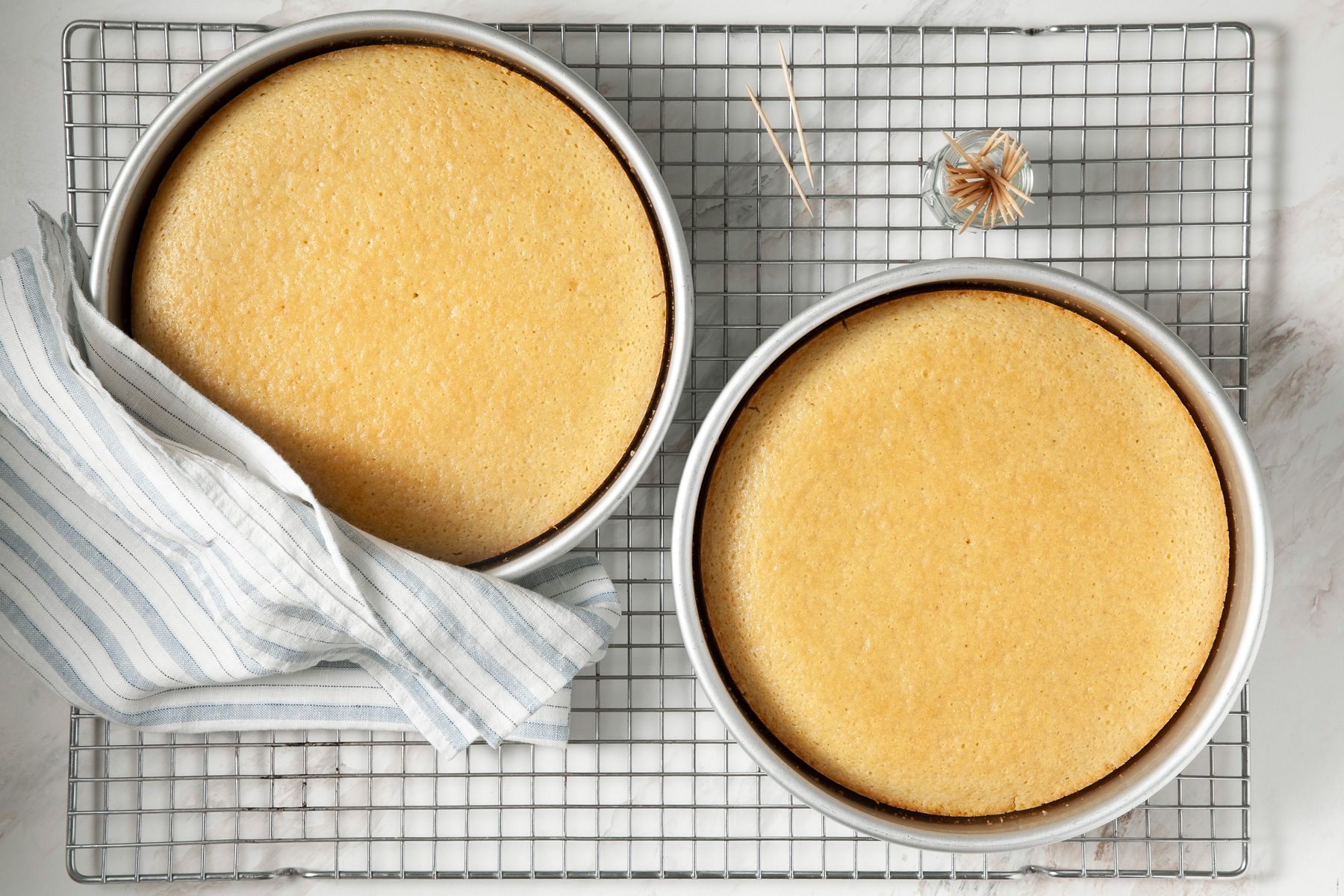 Baked cakes in two pans sit on a wire rack next to toothpicks.