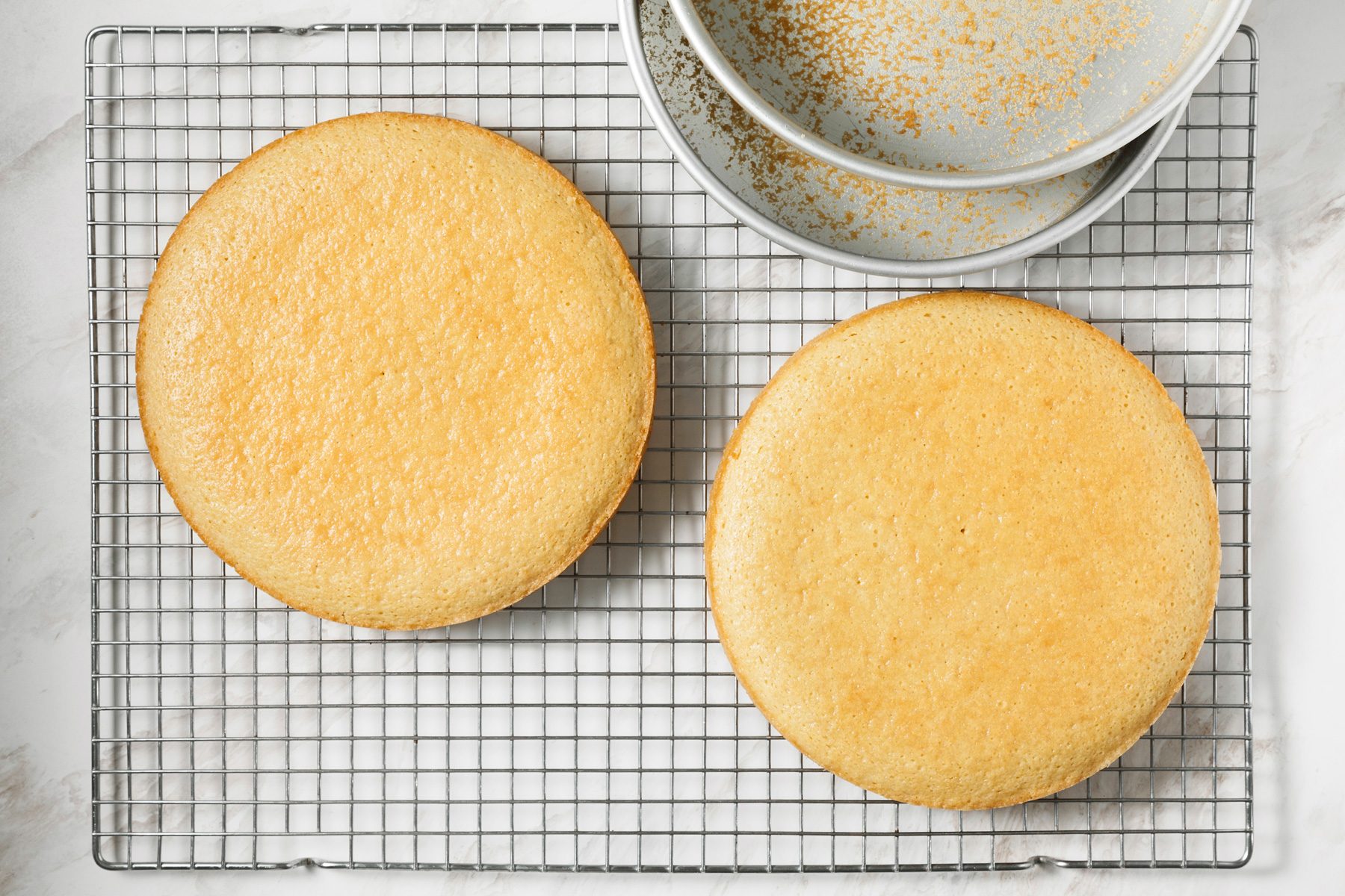 Two baked cakes sit on a wire rack next to pans.