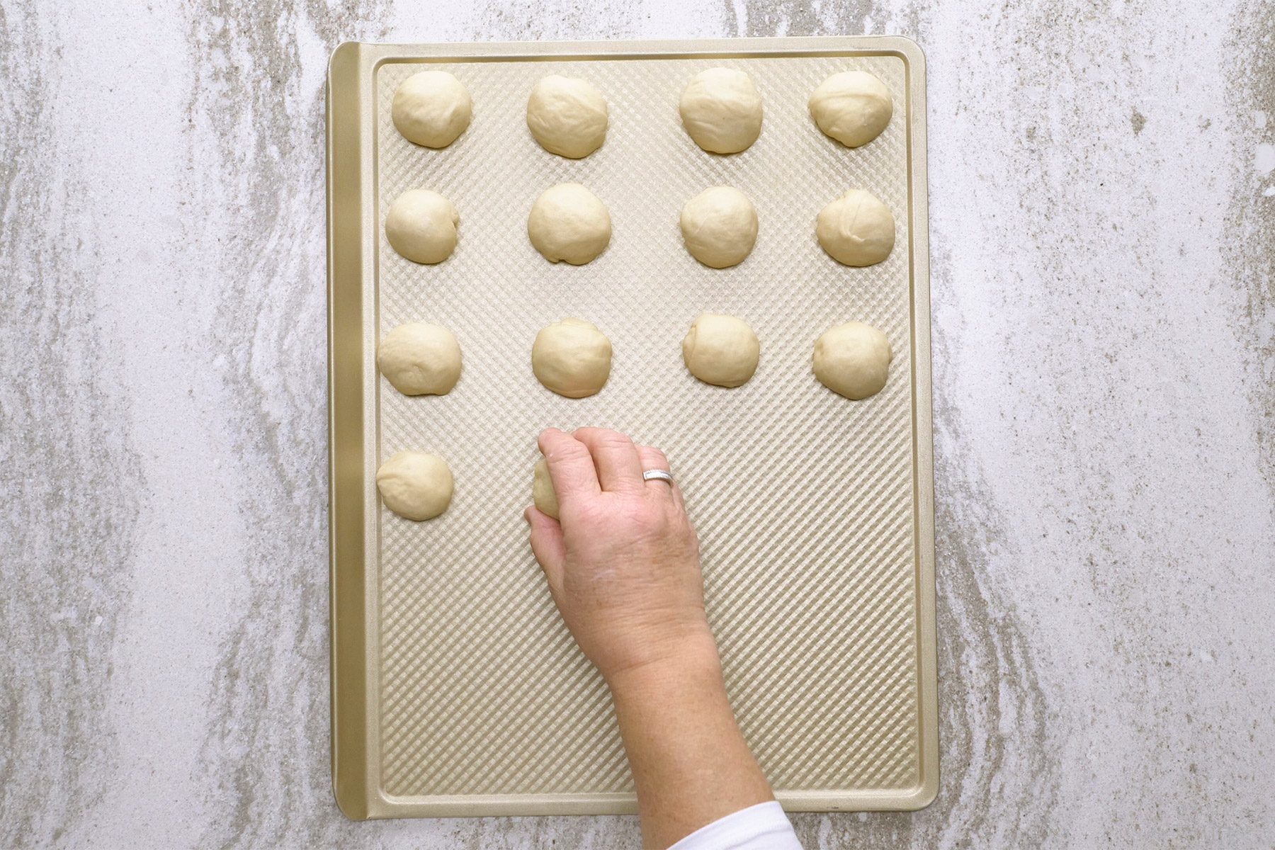A hand places dough balls on a baking sheet arranged in rows, set on a marble countertop.