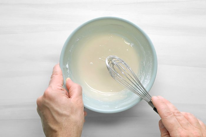 overhead shot of a person is mixing a white liquid in a light blue bowl using a wire whisk; the bowl is on a white table, with a white background;
