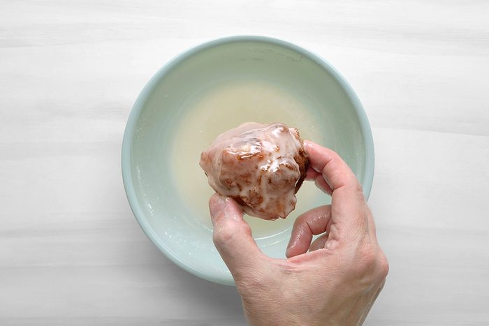 overhead shot of a hand holding a glazed donut that is being dipped into a bowl of glaze; the bowl is light blue and sits on a white surface; the hand is holding the donut by the bottom with the fingers curled around it;