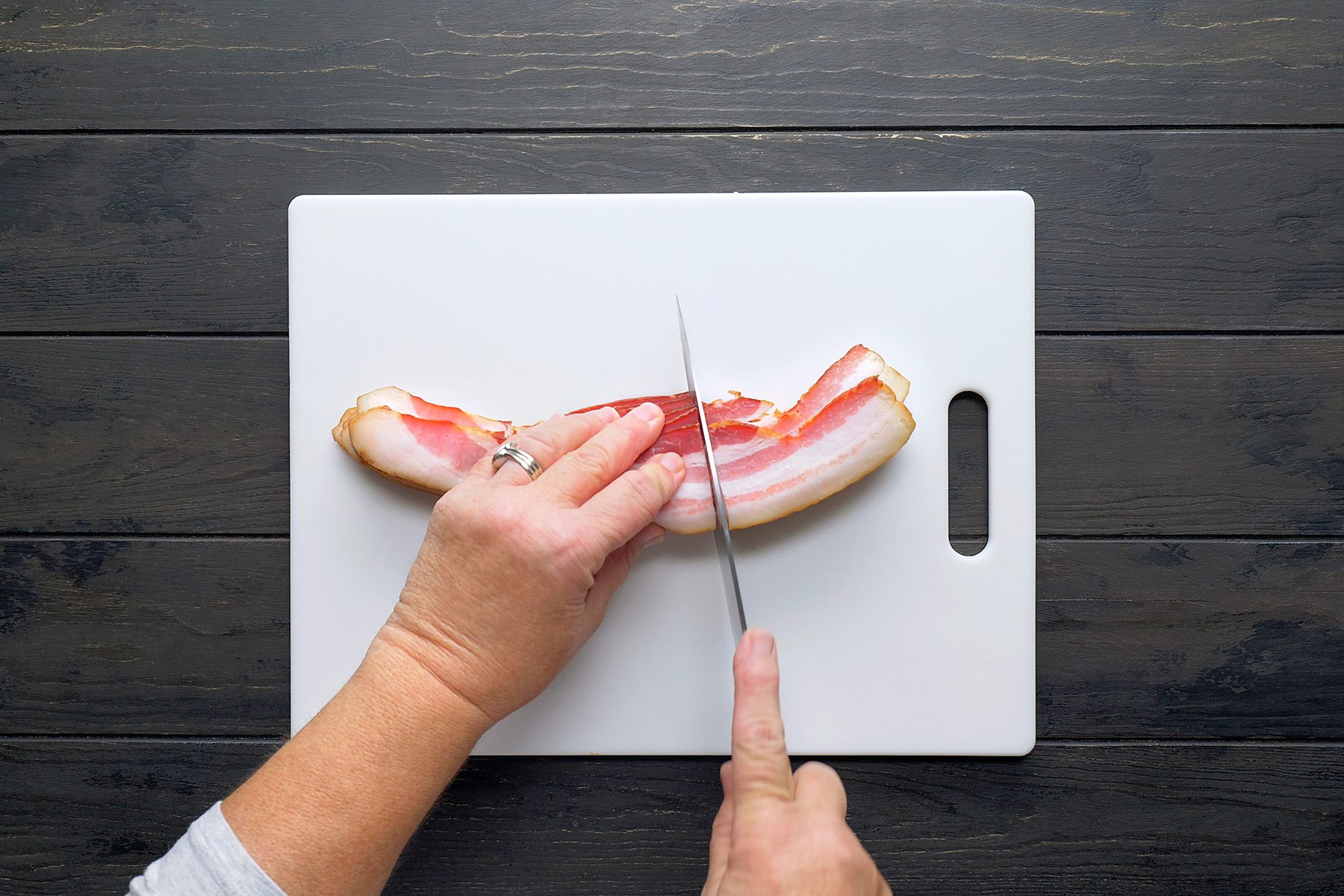 A person slicing raw bacon strips on a white cutting board with a knife. The board is placed on a dark wooden surface. The person is wearing a gray long-sleeve shirt and a ring on their left hand.