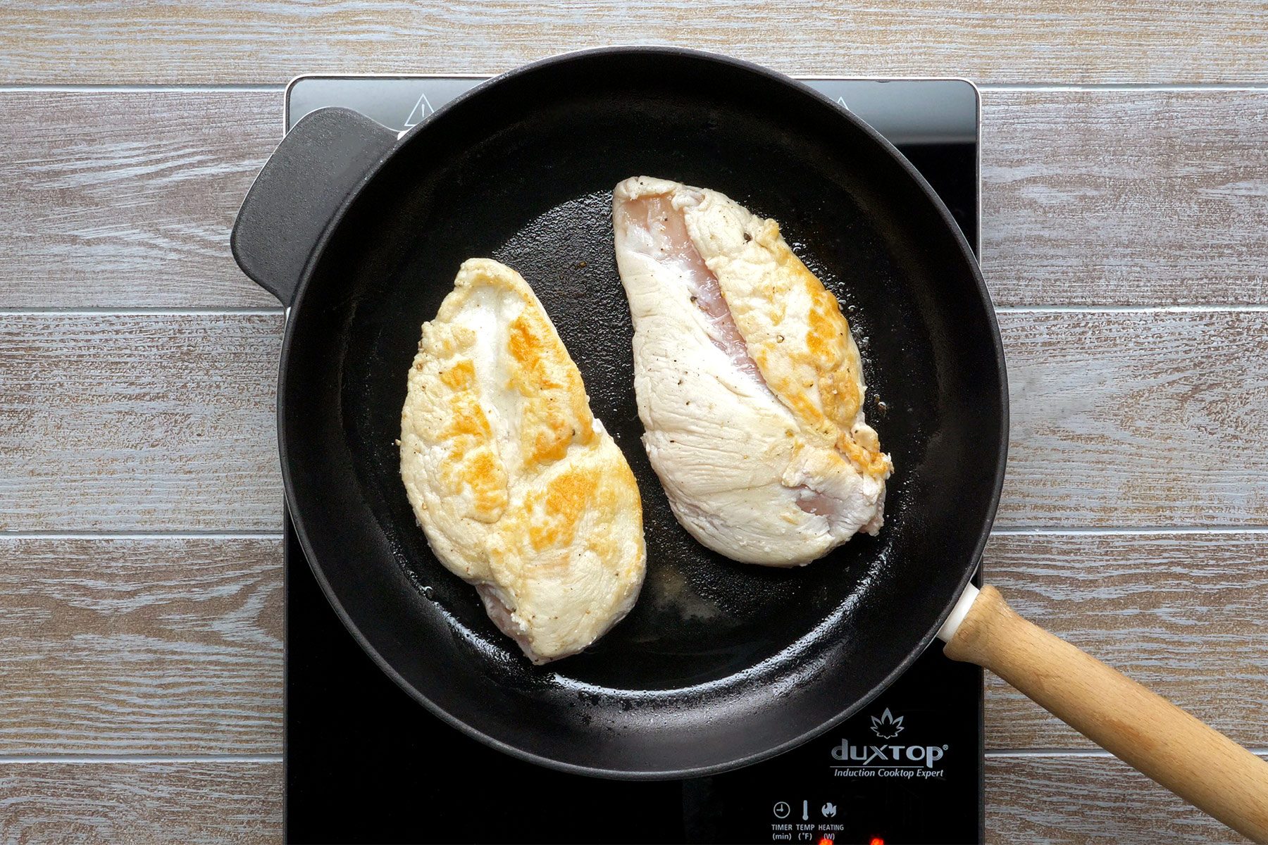 overhead shot of two chicken breasts cooking in a black frying pan on an induction cooktop; the background is made of wood with a light brown stain