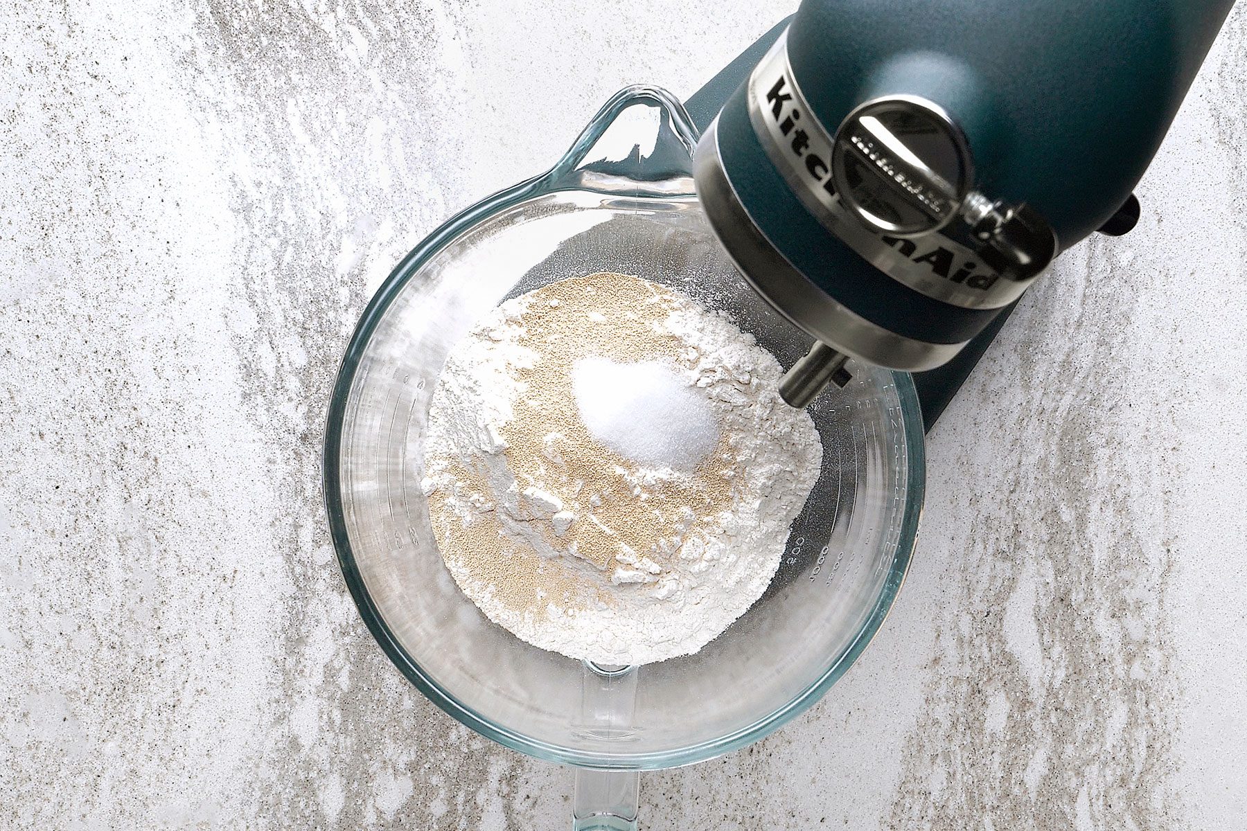 A stand mixer with a glass bowl containing flour and sugar on a textured white and gray surface.