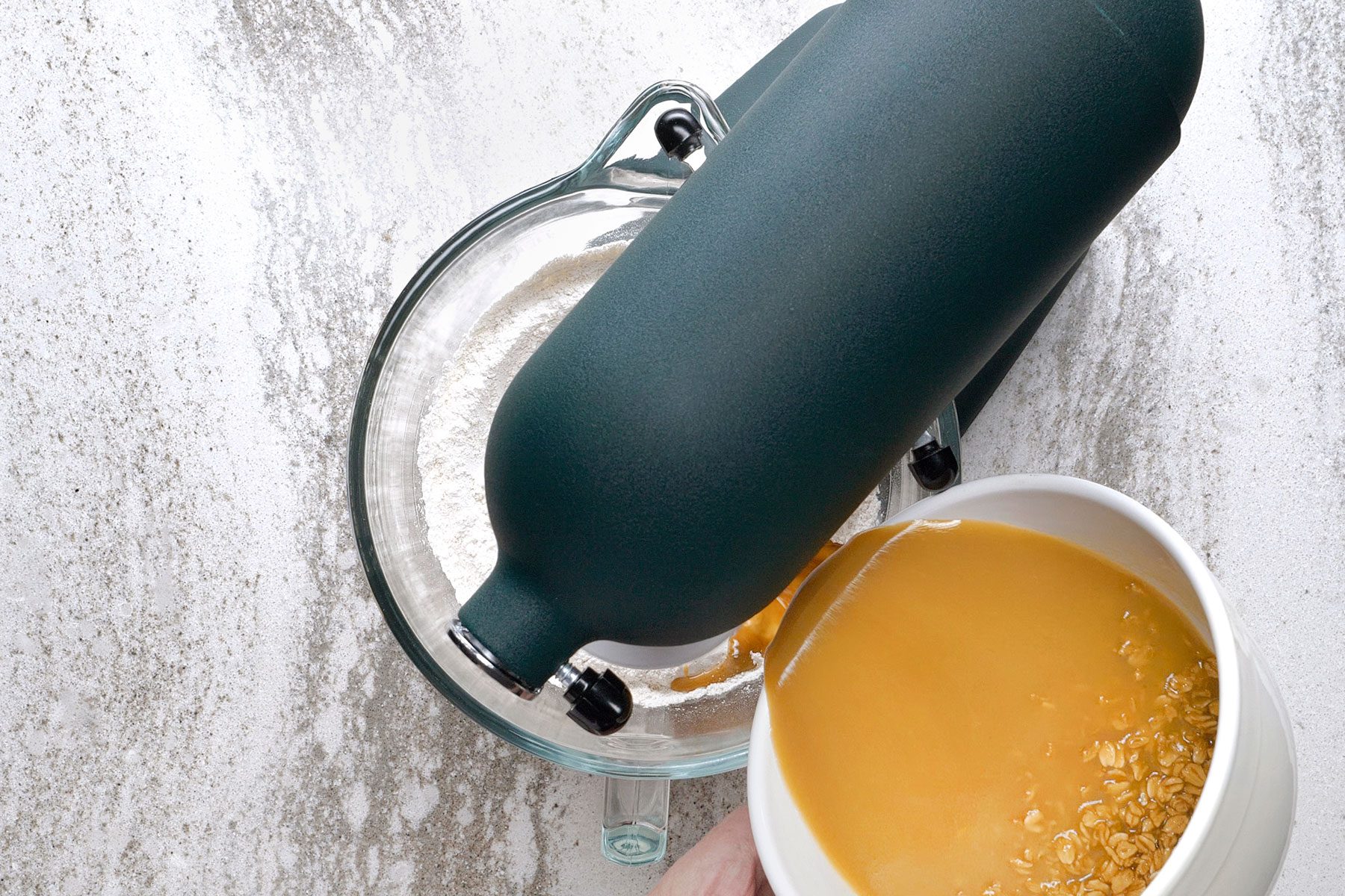 A person pours a bowl of orange liquid and oats into a stand mixer on a speckled countertop. The mixer contains flour.