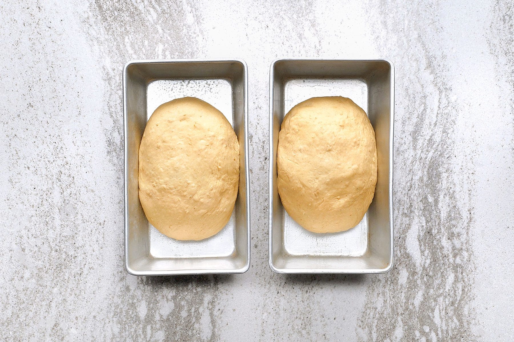 Two rectangular bread pans on a marble surface, each holding a rounded, unbaked dough ball ready for proofing or baking.
