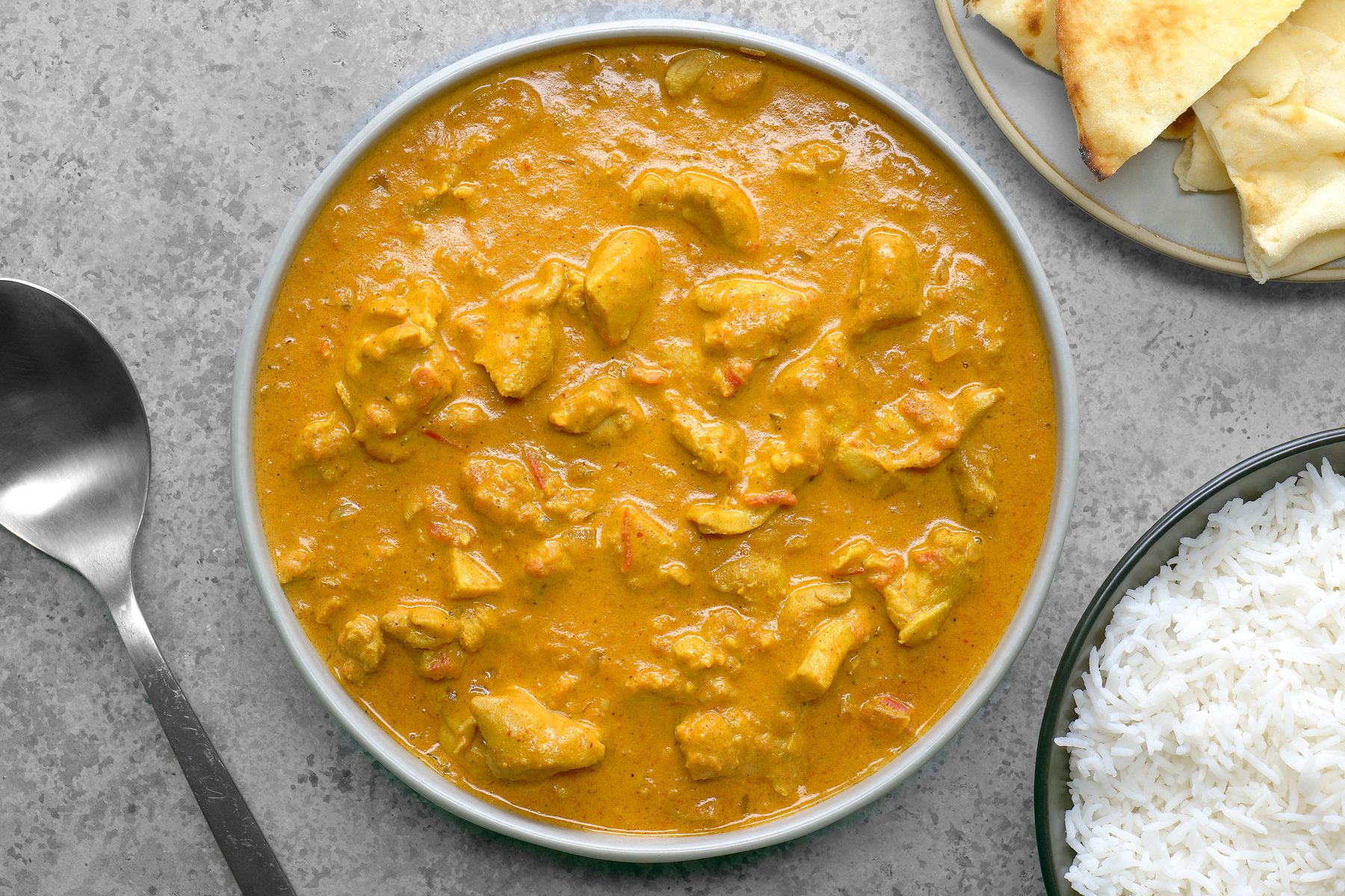 A vibrant serving of butter chicken is presented on a plate, accompanied by fluffy white rice and warm naan, both served on separate plates. The rich, creamy sauce of the butter chicken glistens, inviting a delicious pairing with the side dishes.