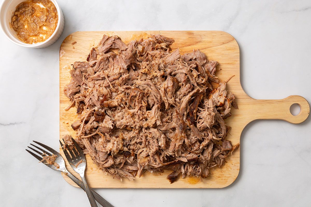 Shredded pork on a cutting board with smashed garlic in a bowl for step two of Chipotle Carnitas recipe for Taste of Home
