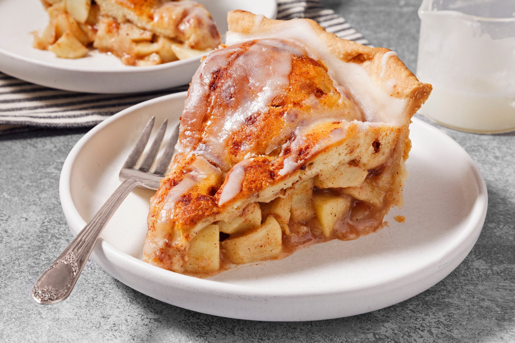 3/4th shot of a slice of apple pie sits on a white plate, with a silver fork placed on the plate, and the handle of the fork is visible; the plate is on a grey textured surface; a blurred view of another slice of apple pie on a white plate can be seen to the left of the main slice; a pitcher of milk is slightly out of focus to the right; the apple pie is topped with a white glaze and cinnamon; the pie crust is a light golden brown