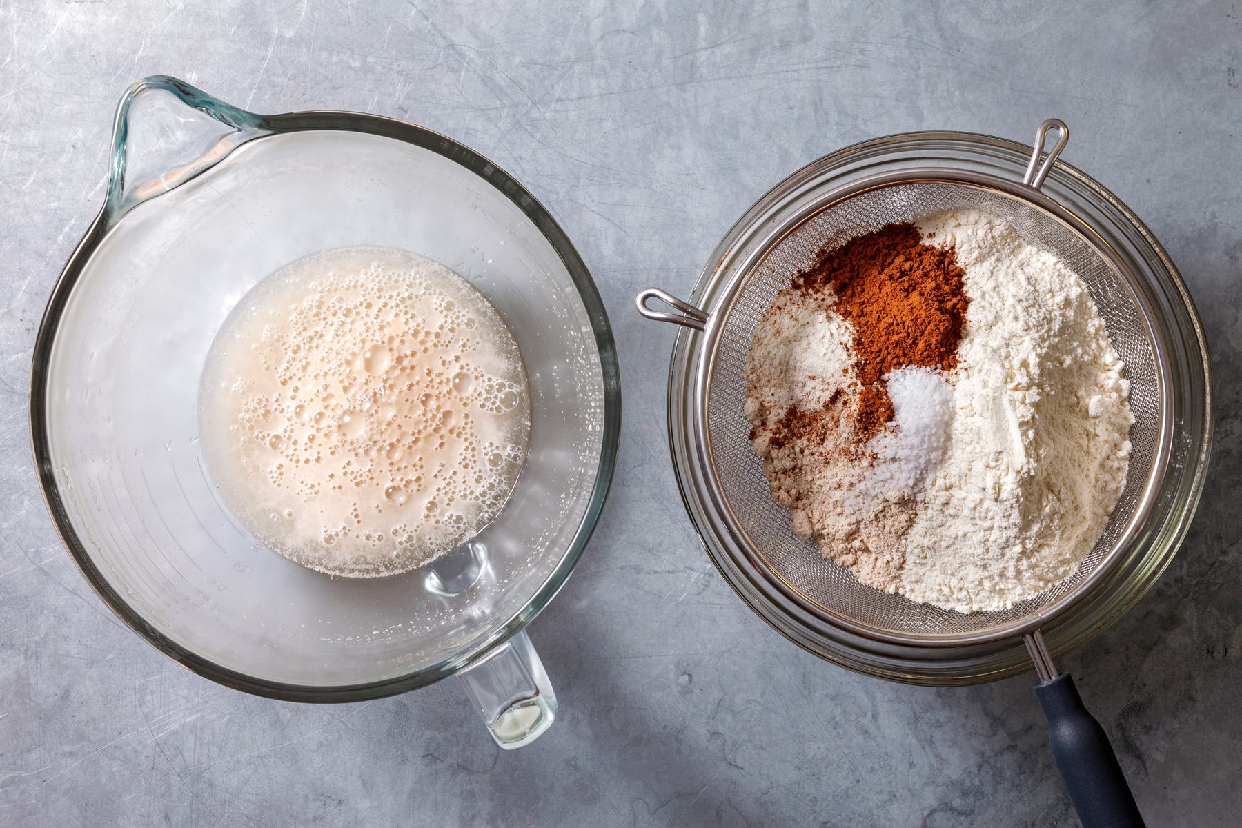 Overhead shot of the bowl of a stand mixer fitted with a dough hook; combine yeast; warm water and sugar; Let sit 5 minutes or until foamy; Meanwhile; in a separate bowl; sift together bread flour; whole wheat flour; cocoa powder and salt; set aside; grey surface;