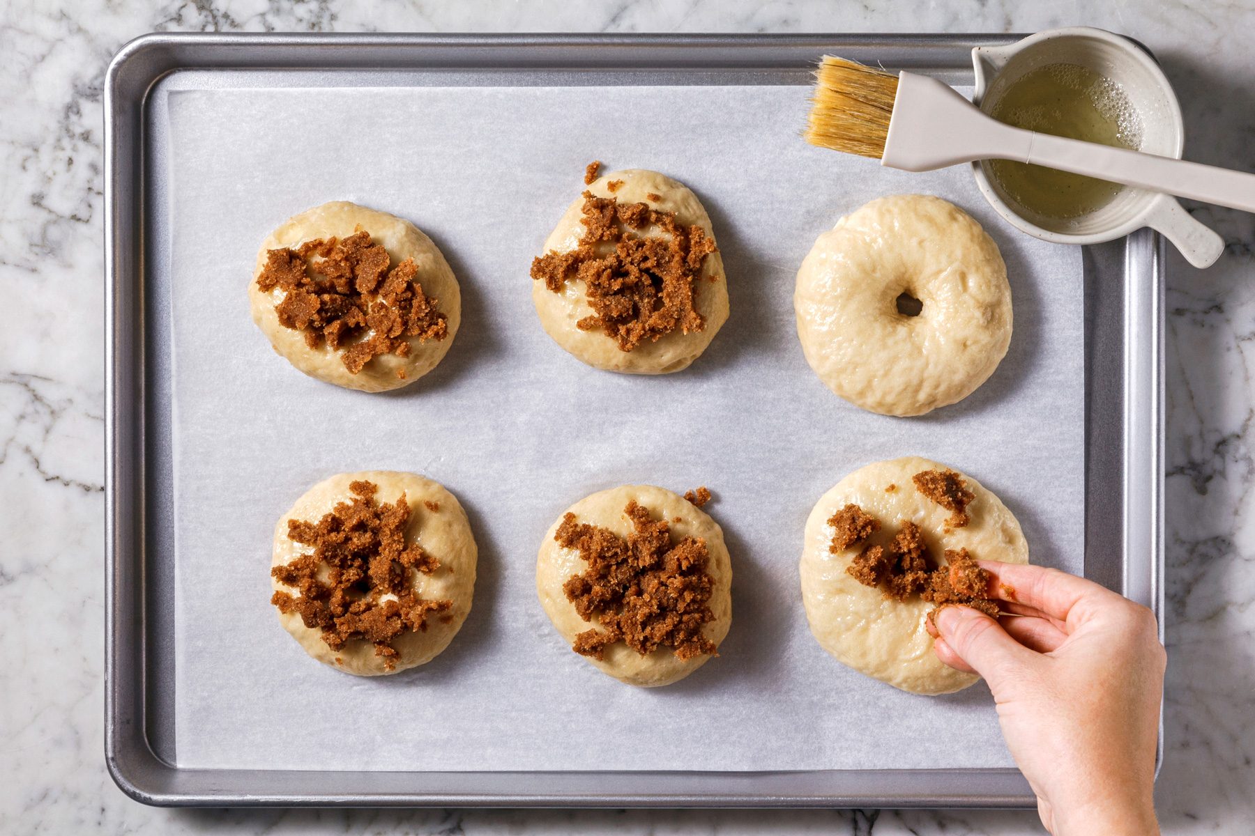 Overhead shot of placing bagels 2 inches apart on greased or parchment-lined baking sheets; brushing with egg white mixture and sprinkling with cinnamon sugar crunch before baking until golden brown, 20-25 minutes, on a marble surface.