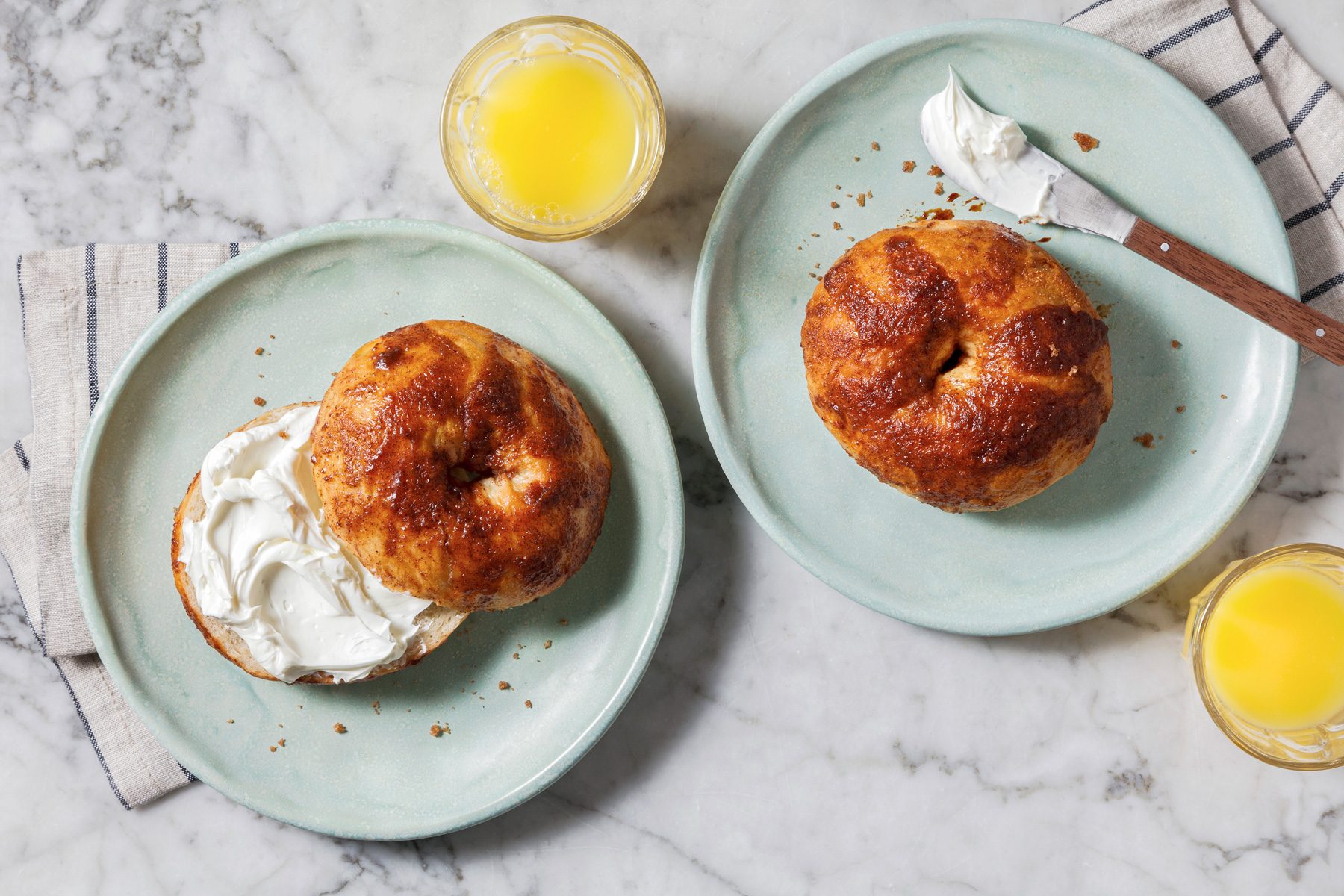 Overhead shot of Copycat Panera Bread Cinnamon Crunch Bagels served on two plates with whipped cream, a knife, drinks, and a napkin on a marble surface.