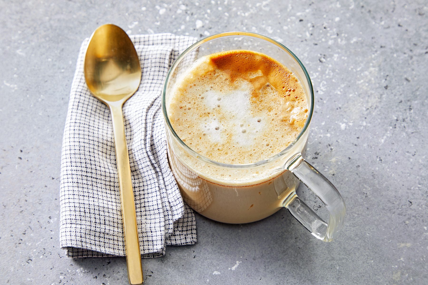 A glass mug of frothy coffee sits on a speckled gray surface next to a folded checkered napkin with a golden spoon on top.