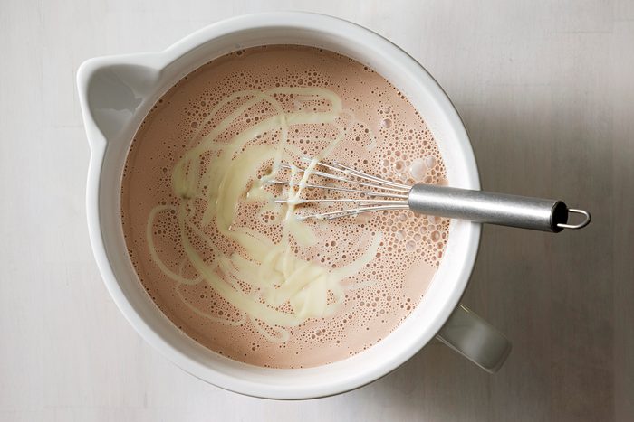 A mug filled with hot chocolate, topped with a swirl of cream. A metal whisk rests inside the mug, indicating the drink has just been stirred. The mug sits on a light-colored surface.