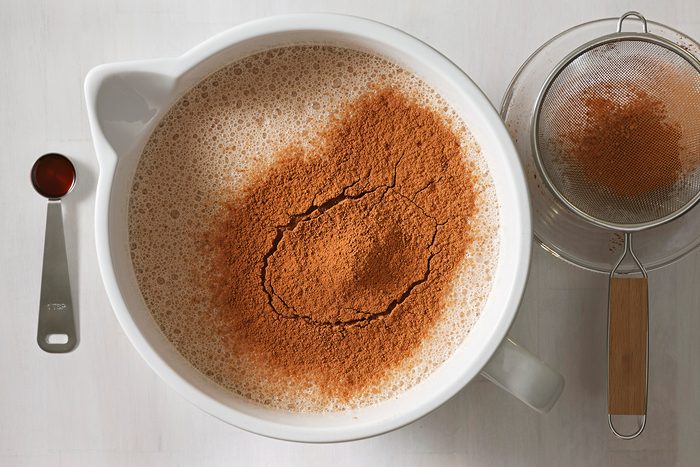 A mixing bowl filled with liquid and a heap of cocoa powder on top, next to a teaspoon of vanilla extract and a fine mesh sieve with cocoa residue on a light gray surface.
