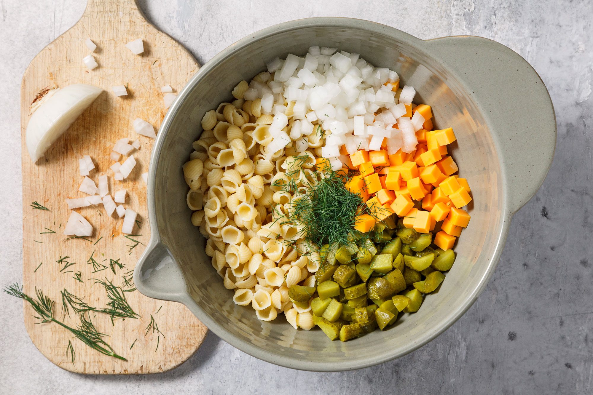 Overhead shot of a large bowl; combine pasta; pickles; cheese; onion and dill; wooden board; grey surface;