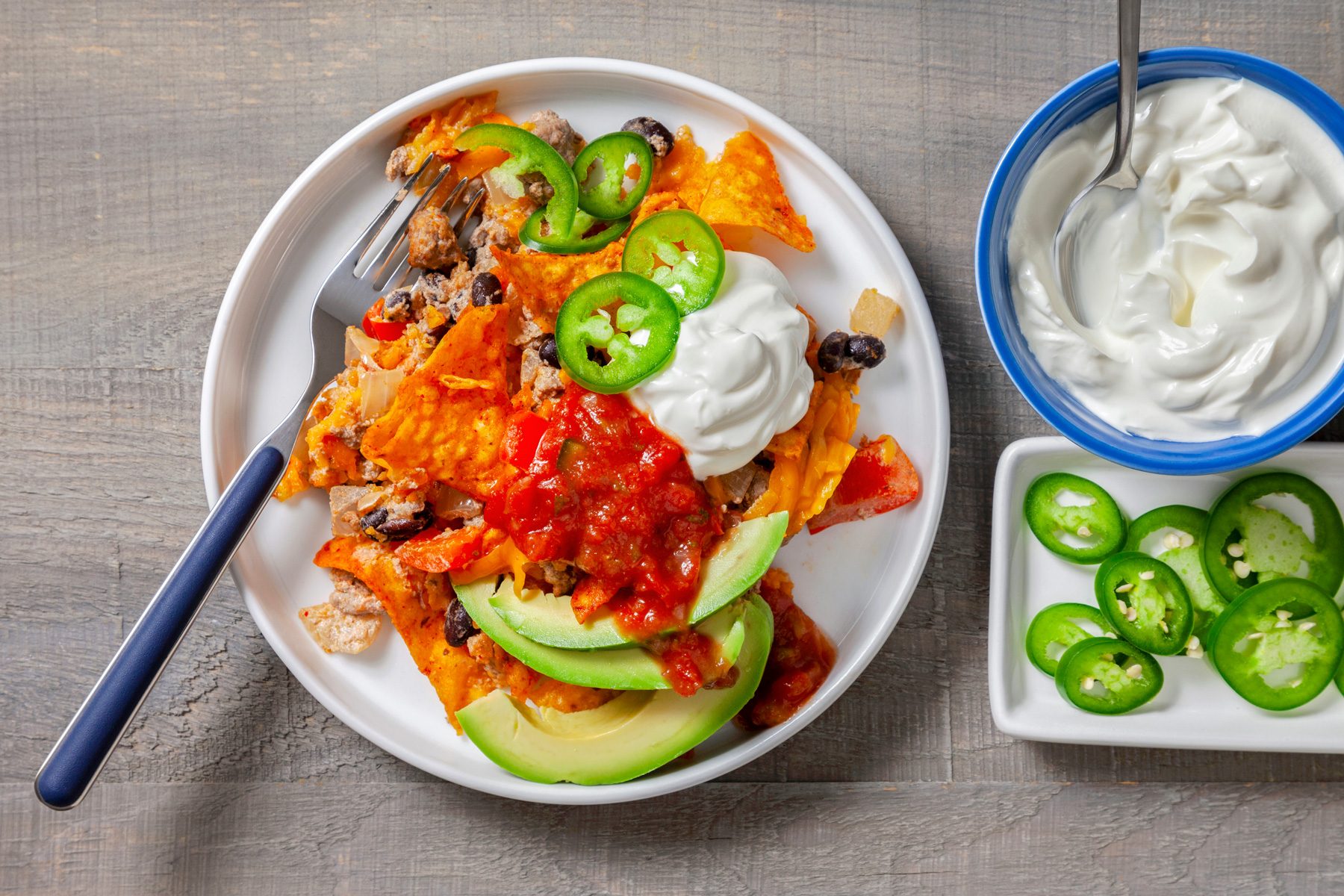 Overhead view of a Doritos casserole topped with sliced peppers and served with a side of sour cream.