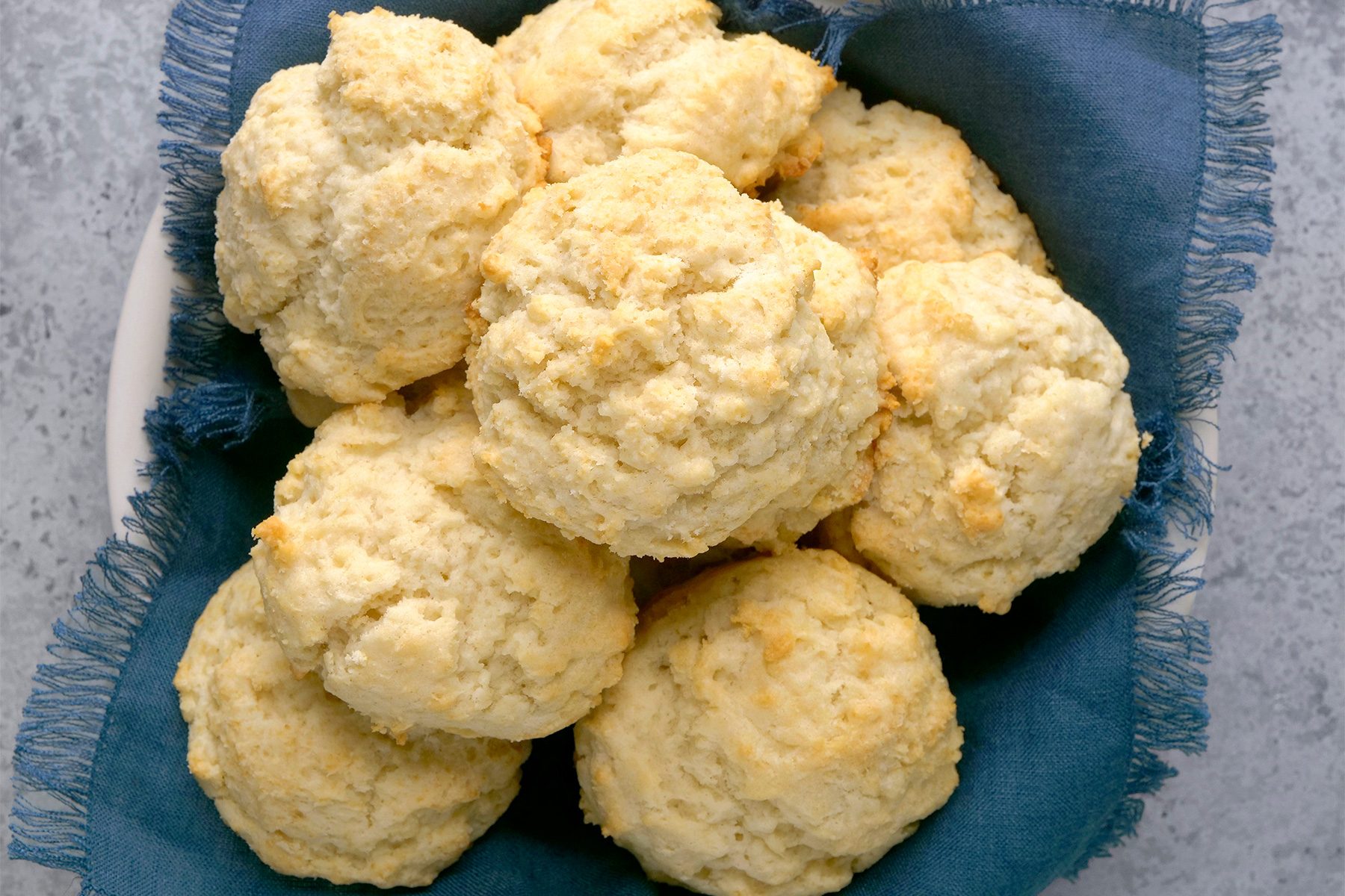 A basket filled with freshly baked biscuits placed on a dark blue cloth. The biscuits have a golden-brown, crumbly texture, indicating they are homemade and soft. The background is a light gray surface.
