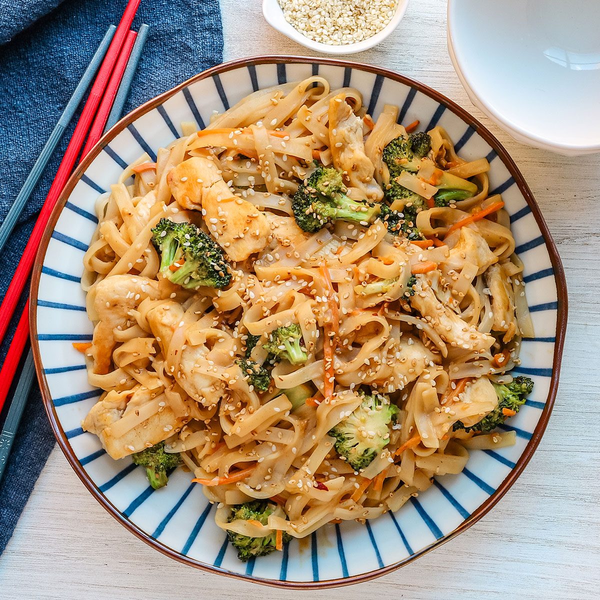 A serving bowl of peanut chicken stir-fry with broccoli, shredded carrots, sesame seeds and an Asian peanut sauce.
