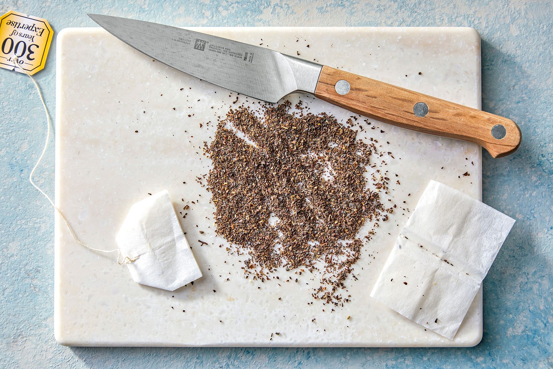 A wooden-handled knife rests on a marble cutting board next to loose black tea leaves. Two tea bags, one opened, and a paper tag with "3000" printed on it are also visible on the board.