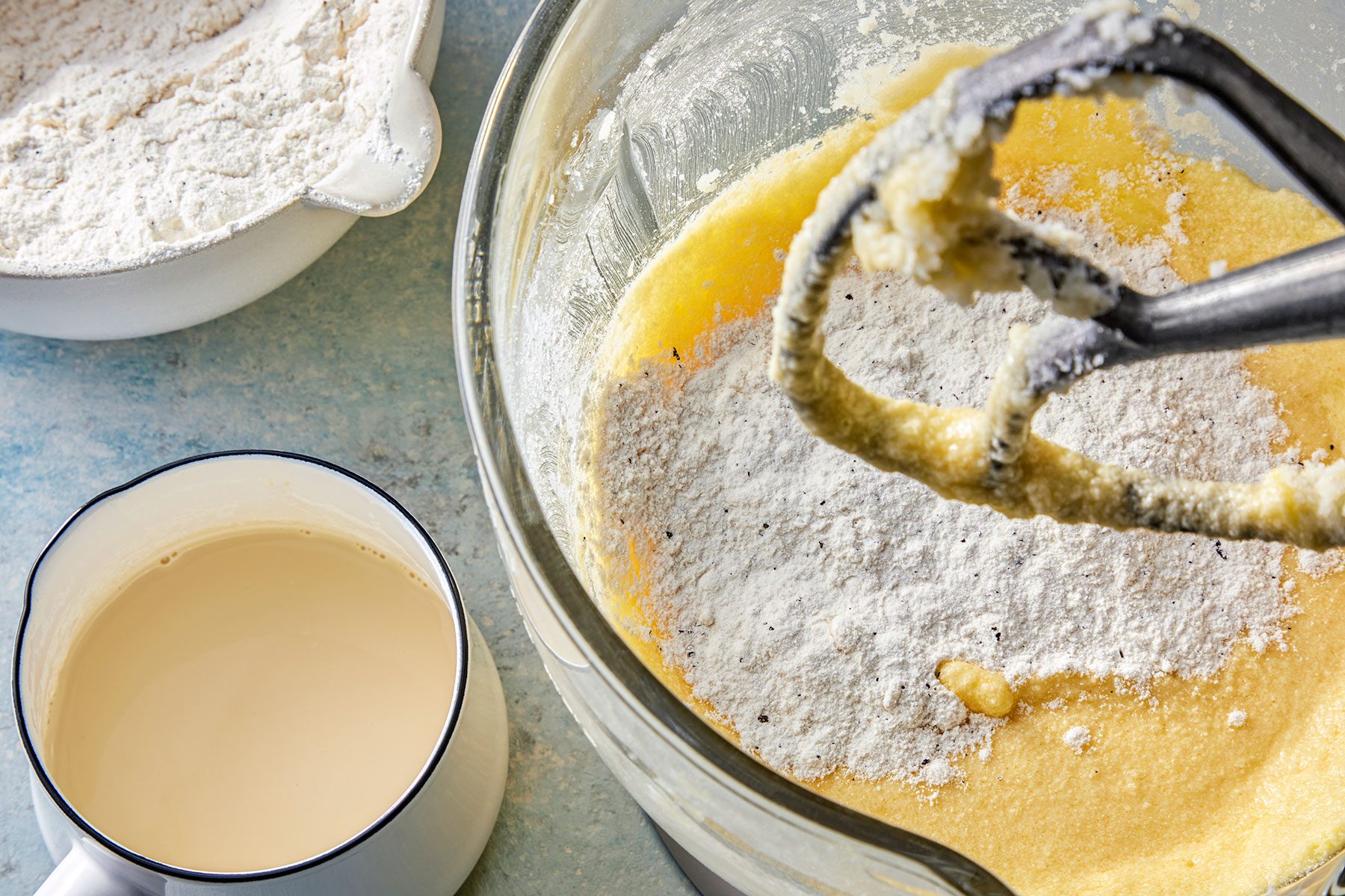 A mixing bowl contains creamed butter and sugar with flour being incorporated. Next to it are a bowl with flour and a jug of milk, set on a light blue countertop. A mixer attachment is partially visible.