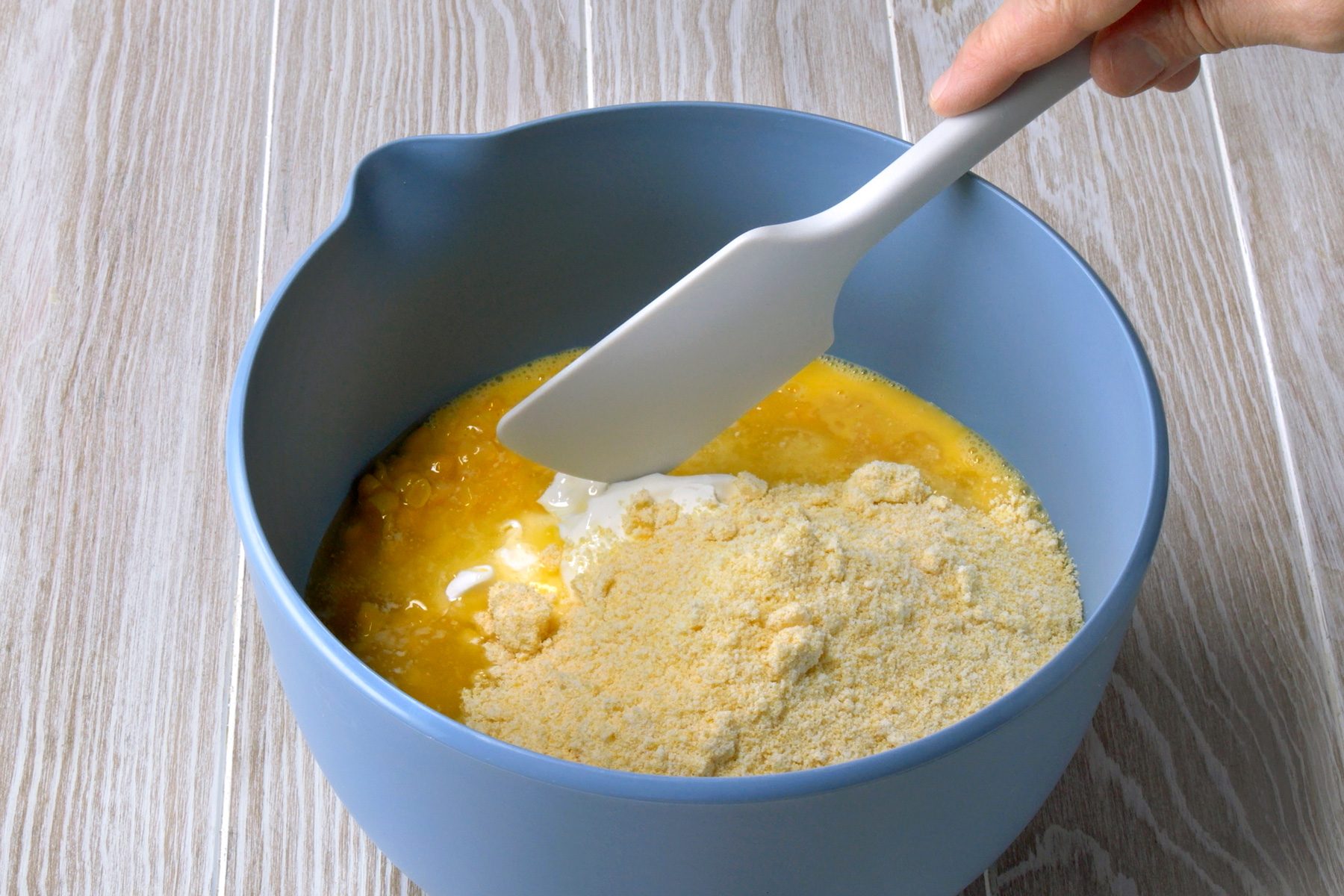 3/4th shot of a blue mixing bowl on a wooden surface, containing a mixture of ingredients for a recipe; the bowl is filled with yellow beaten eggs, a white spatula, held by a hand, is used to combine the ingredients