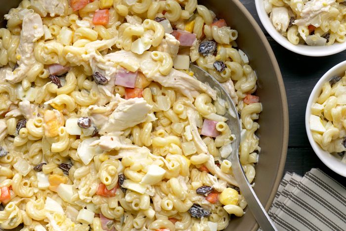overhead shot of a bowl of macaroni salad with a fork in it; the salad is sitting on a brown and white striped napkin; there are two other bowls filled with the same salad on a dark wood table