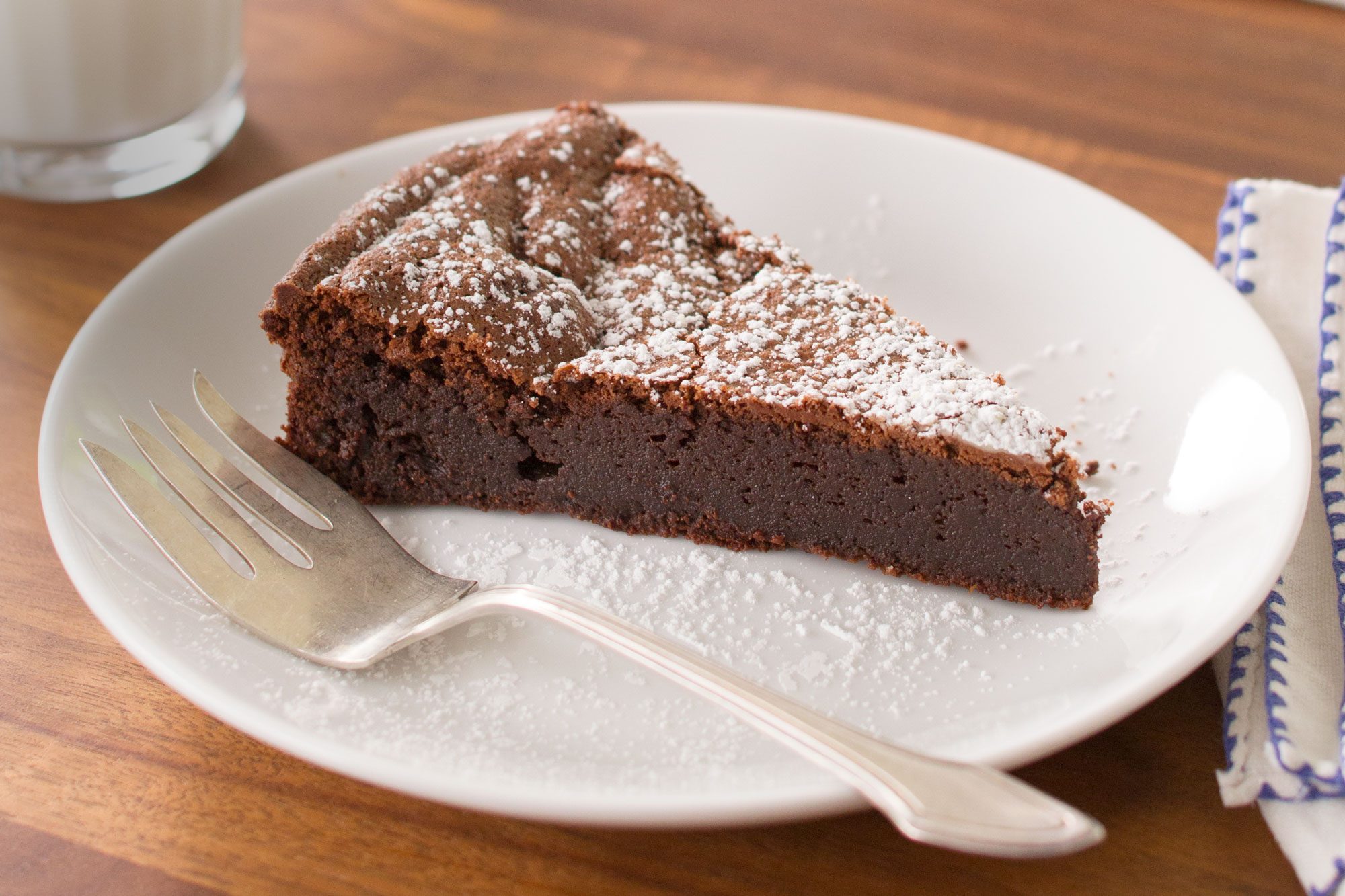 A slice of Flourless Chocolate Torte dusted with powdered sugar sits on a white plate with a fork beside it. The plate is placed on a wooden table next to a blue-striped napkin.