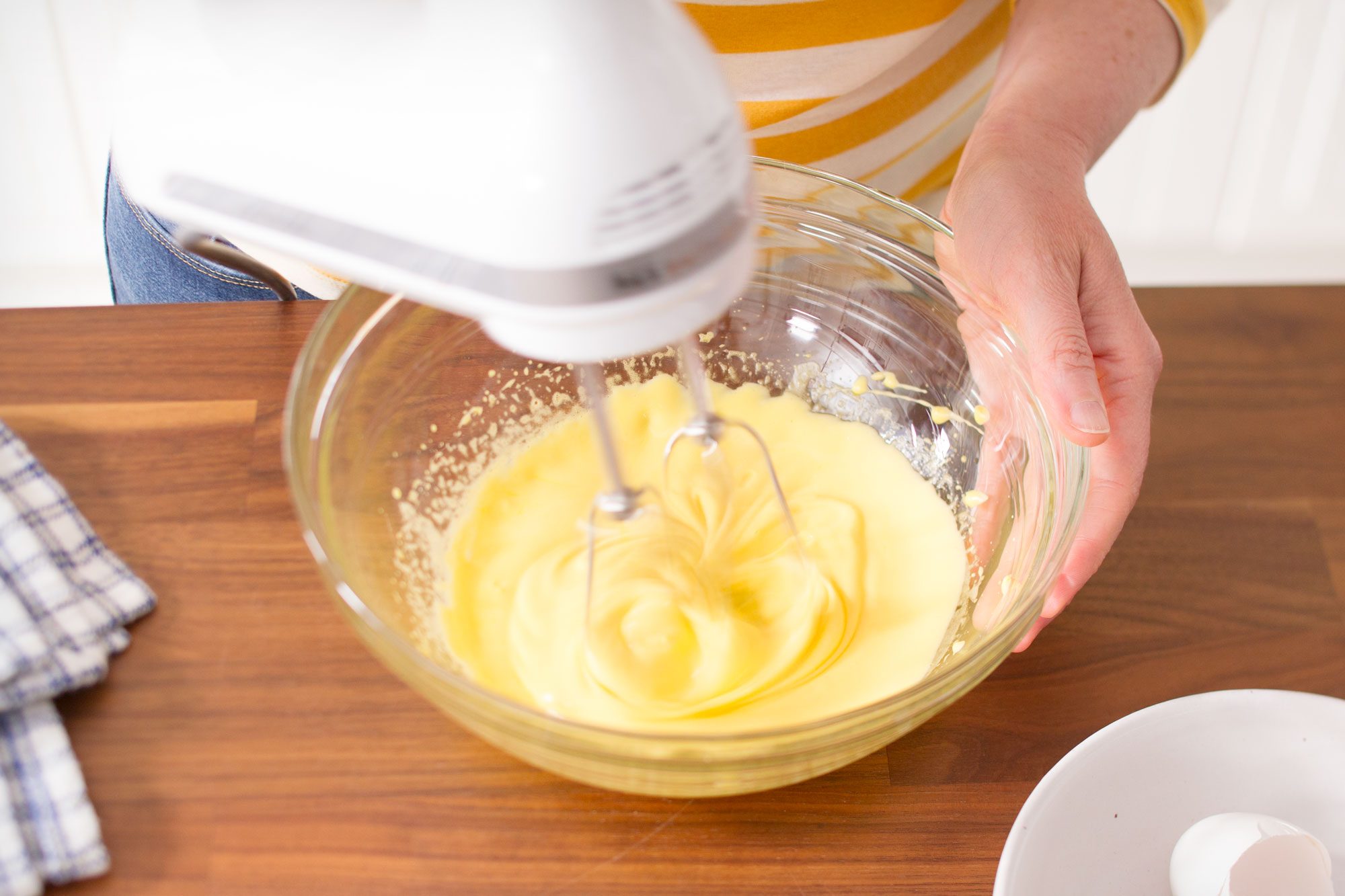A person using a white electric mixer to blend a yellow batter in a clear glass bowl. A cracked egg and a checkered cloth are nearby on a wooden countertop.