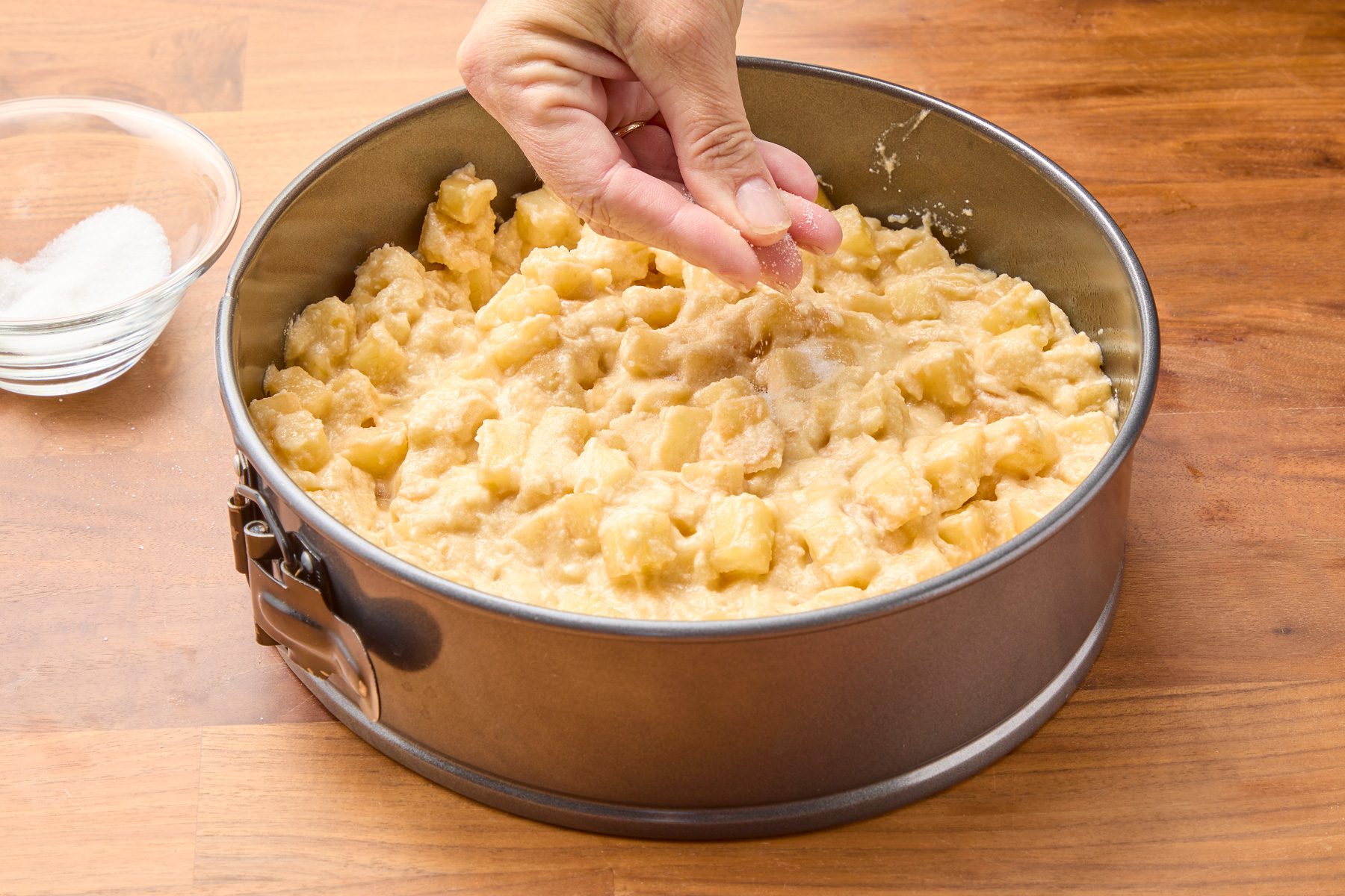 Three-quarter shot of a hand sprinkling sugar onto cake batter in a springform pan. The batter, made with cubed apples, is sitting on a wooden counter, with a small bowl of sugar visible to the left of the pan.