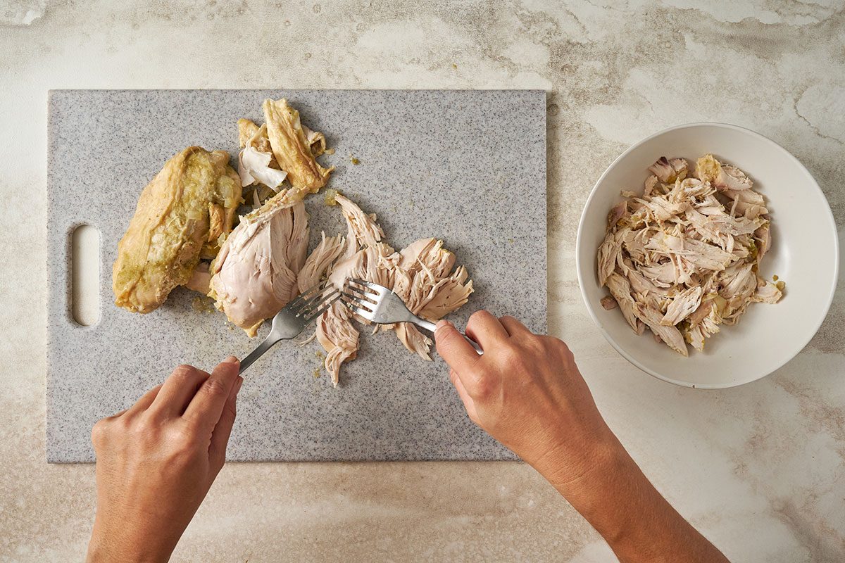 Overhead view of chicken breasts removed from the slow cooker and being shredded with two forks for the Taste of Home Green Chicken Chili recipe.