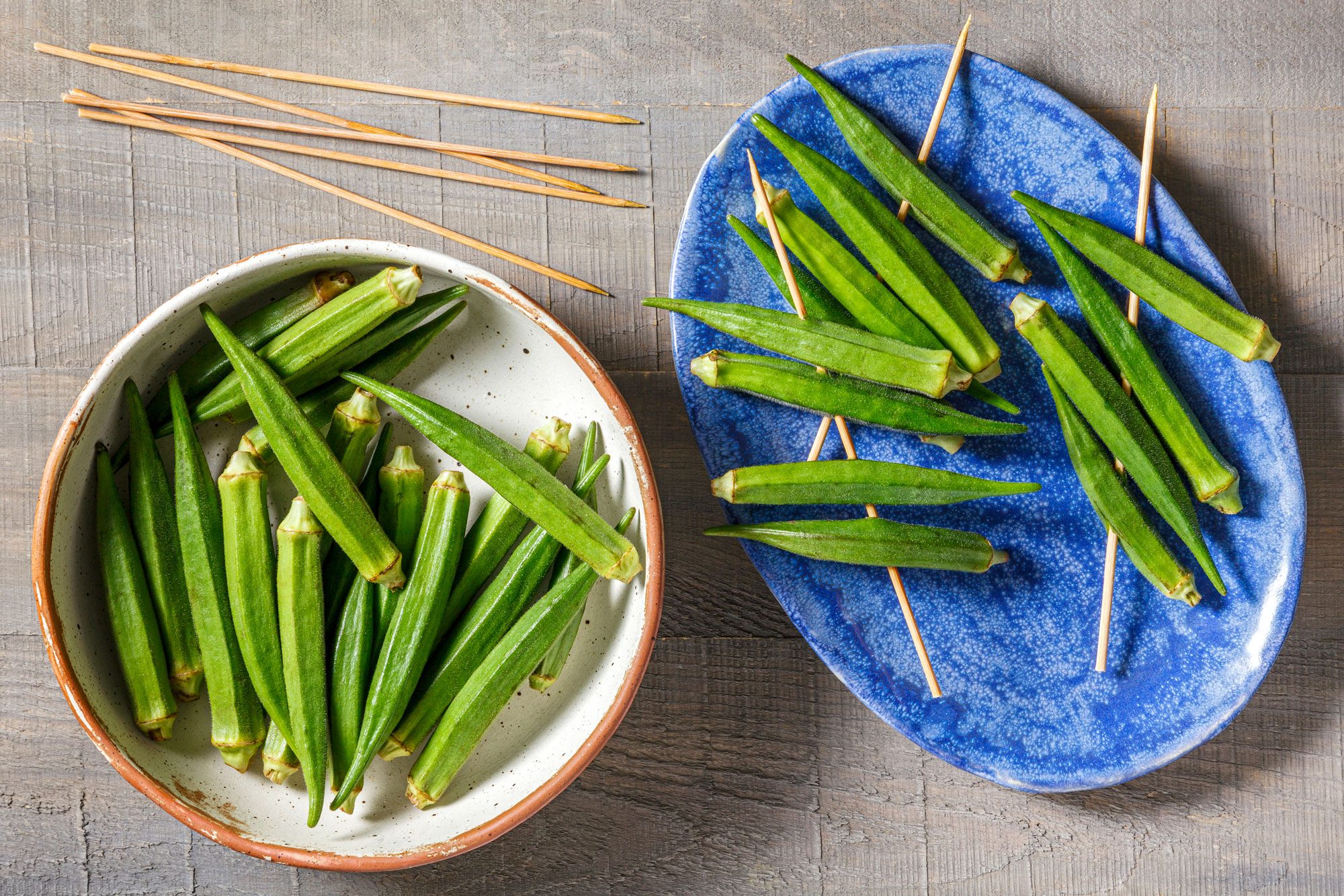 Overhead shot of thread 4-5 okra pods onto each wooden skewer; wooden surface;