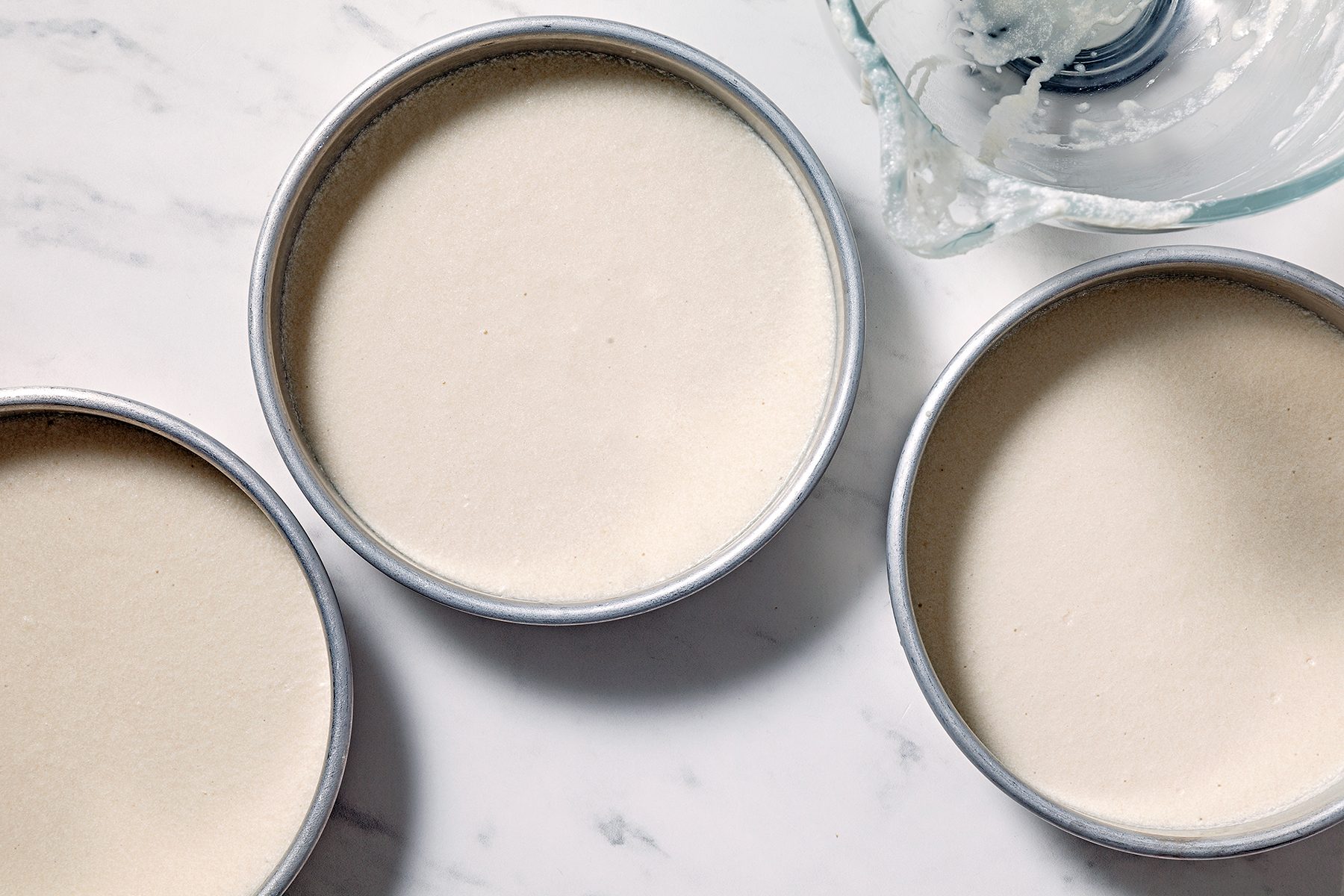 Three round cake pans filled with batter are placed on a marble surface. A glass mixing bowl with some remaining batter is partially visible in the corner.