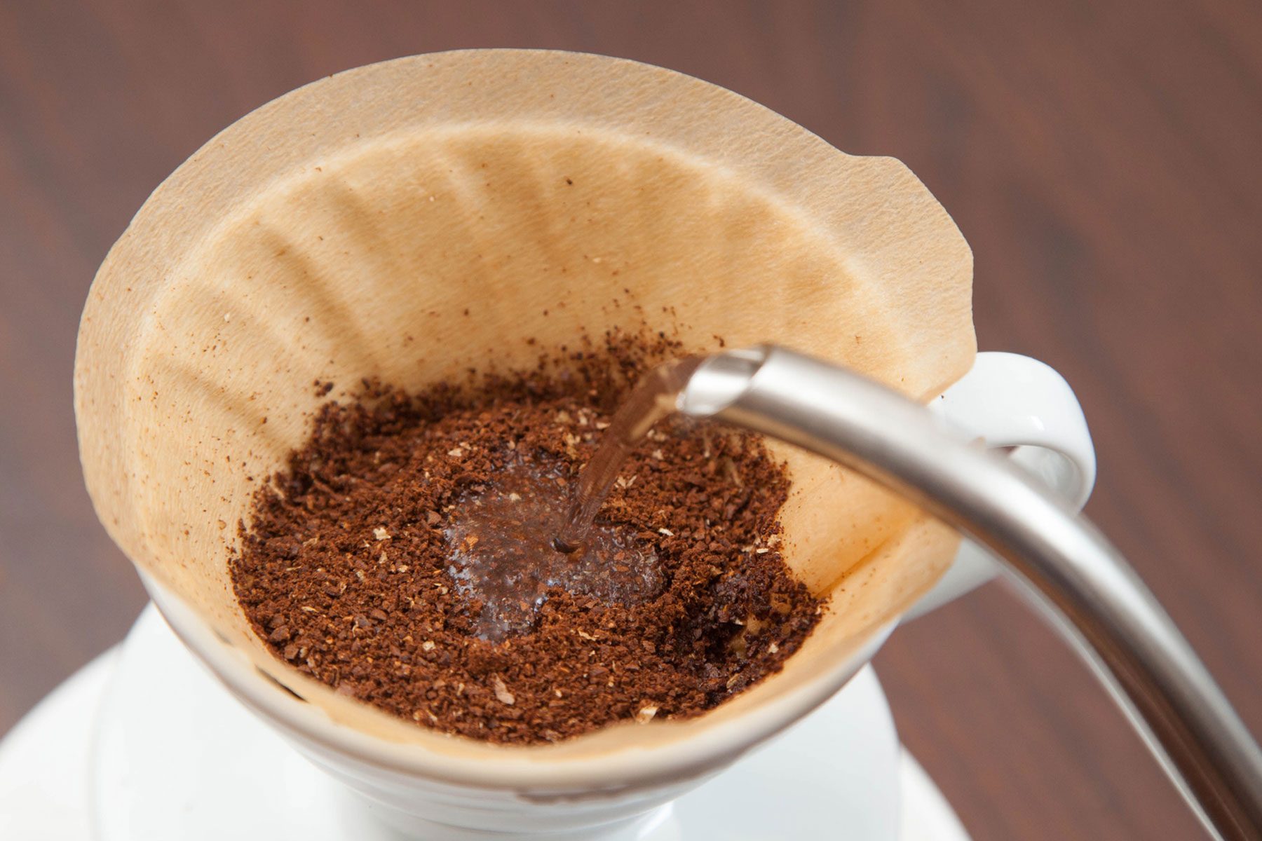Hot water from a pour-over kettle is being poured over coarse ground coffee. The ground coffee sits in brown filter paper over a white drip coffee cone.