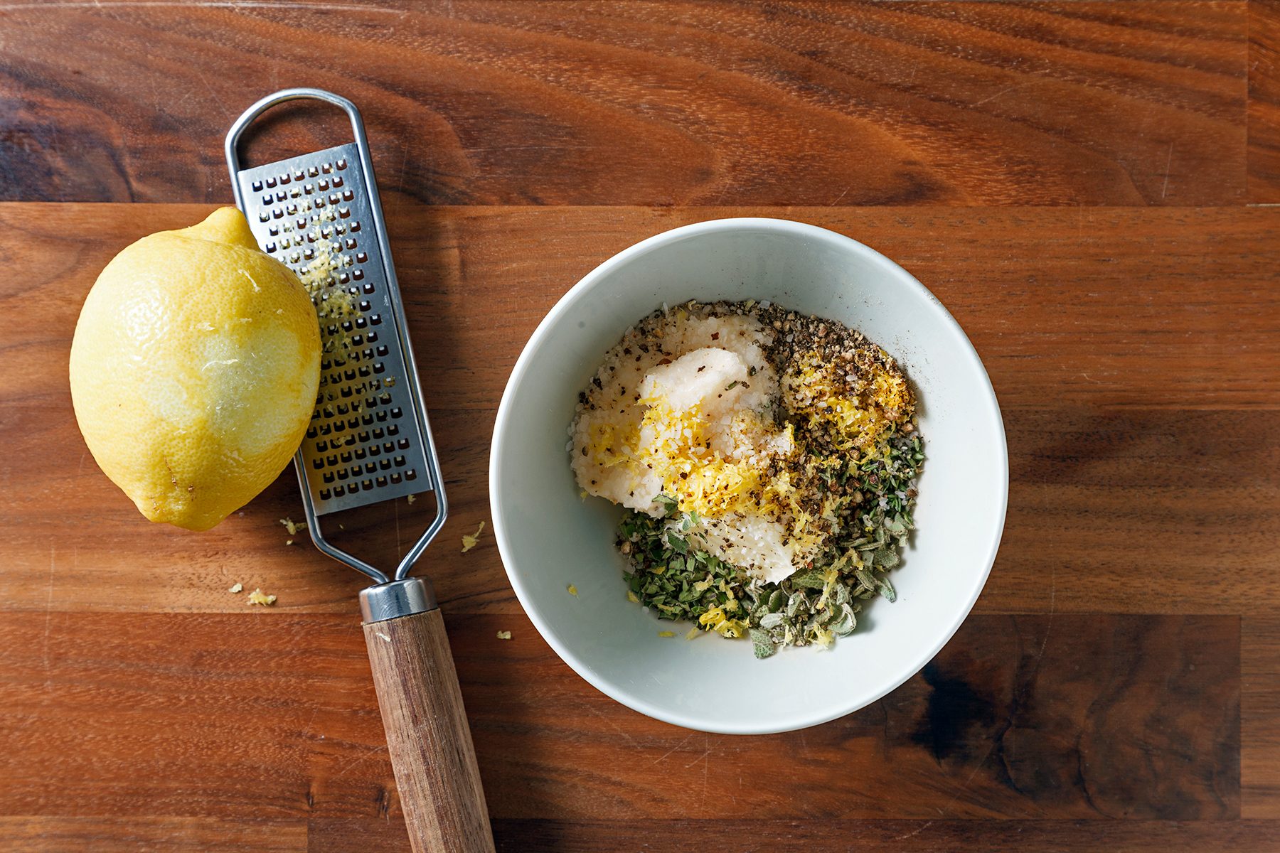 A bowl filled with various spices and lemon zest on a wooden surface. A lemon and a grater are beside the bowl.