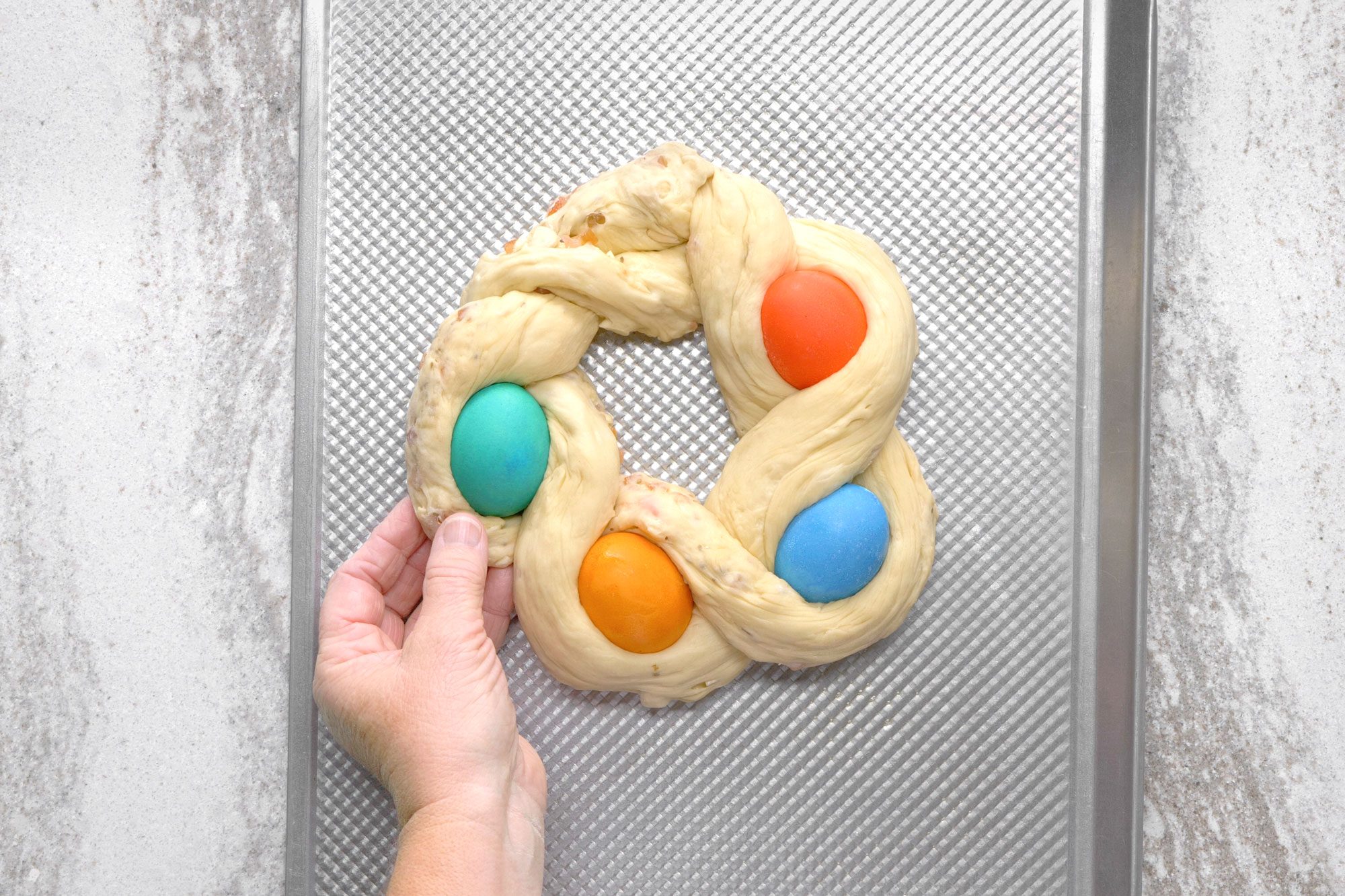 overhead shot of A hand is placing a blue Easter egg into a braided bread dough, on a baking sheet; the dough is in a wreath shape and already contains two eggs, one orange and one blue; the baking sheet is silver and has a diamond pattern; the background is a white concrete surface