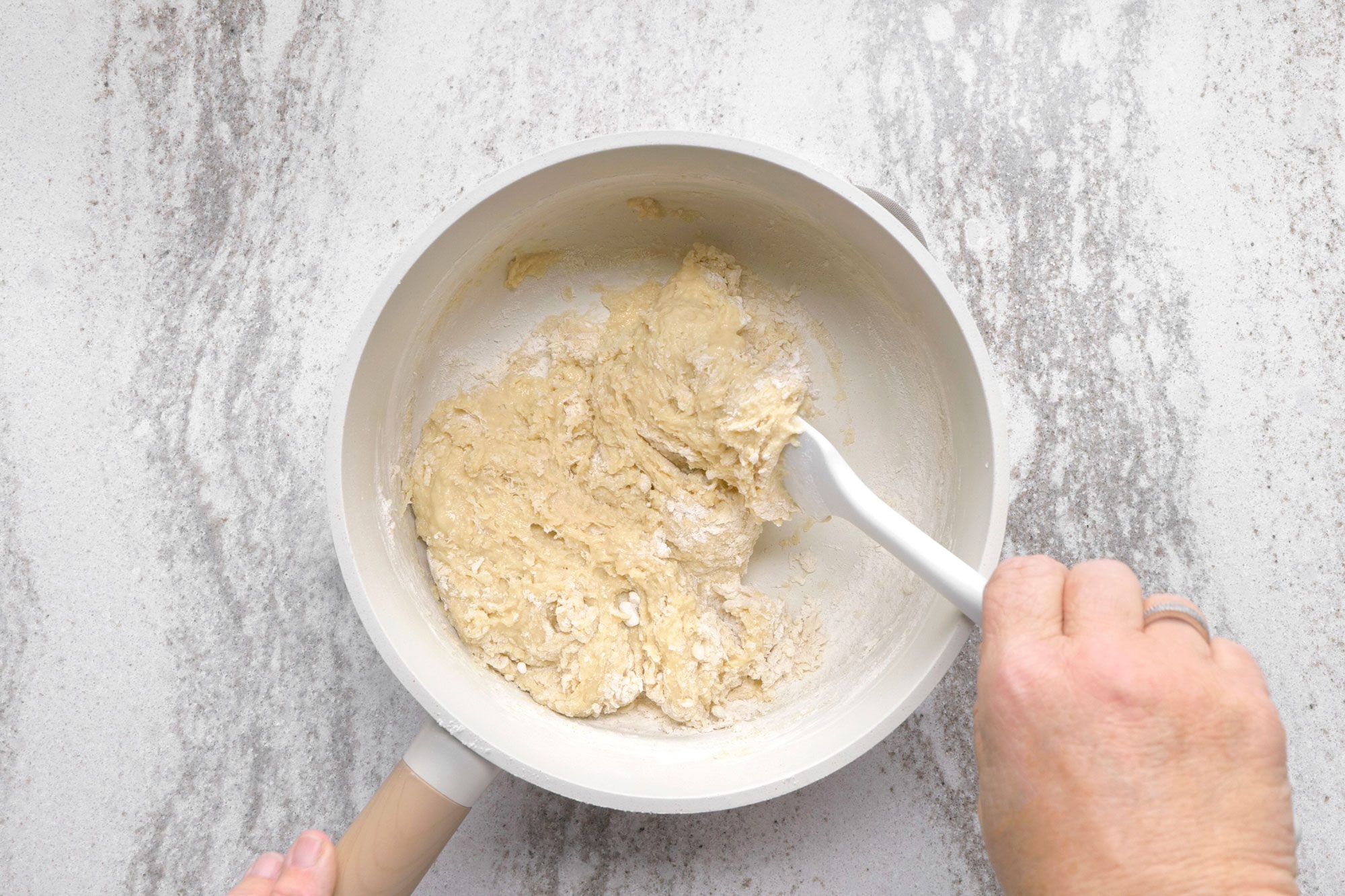 overhead shot of a white bowl with a wooden handle contains dough being stirred by a white spatula; the bowl is on a white countertop with grey speckles, a hand holds the spatula and another hand is to the left of the bowl; the dough looks sticky and soft