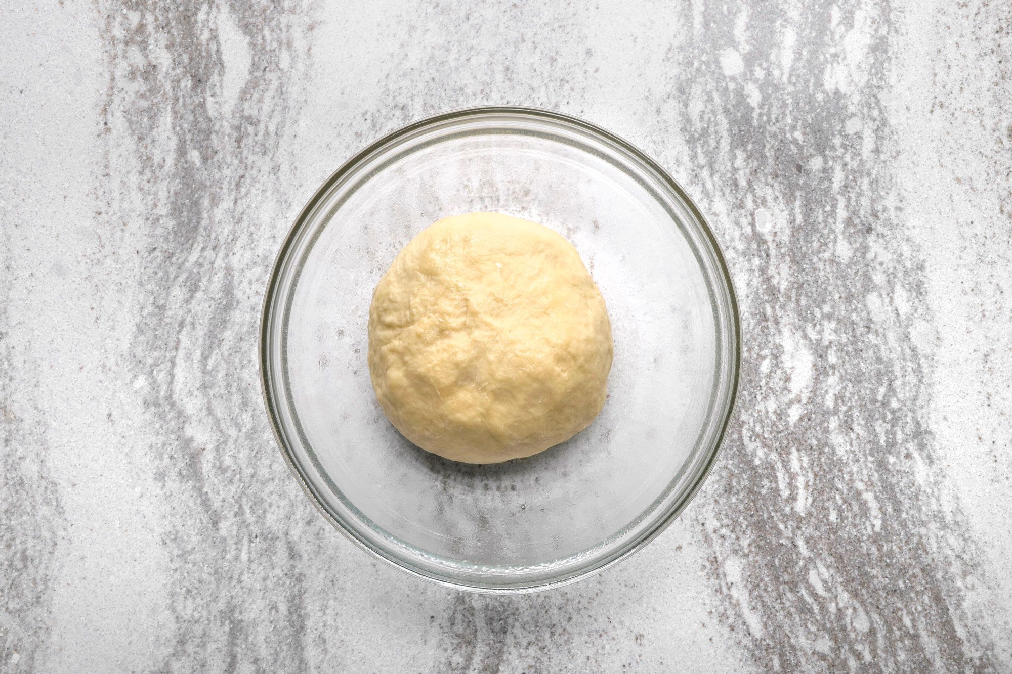 overhead shot of a ball of dough sits in a glass bowl on a white countertop with gray flecks;