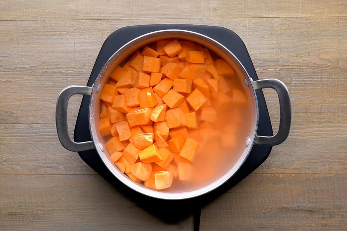 overhead shot of a pot of cubed sweet potatoes submerged in water on a wooden surface; the pot is made of stainless steel with black handles and is sitting on a black heat-resistant pad