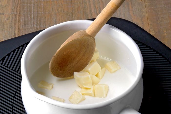 3/4th shot of a white ceramic bowl with a wooden spoon is on top of a black cooking mat, and a wooden table is in the background the bowl contains liquid and pieces of white butter; the spoon is sticking out of the bowl the spoon's handle is also made of wood; the background of the image is a wooden table