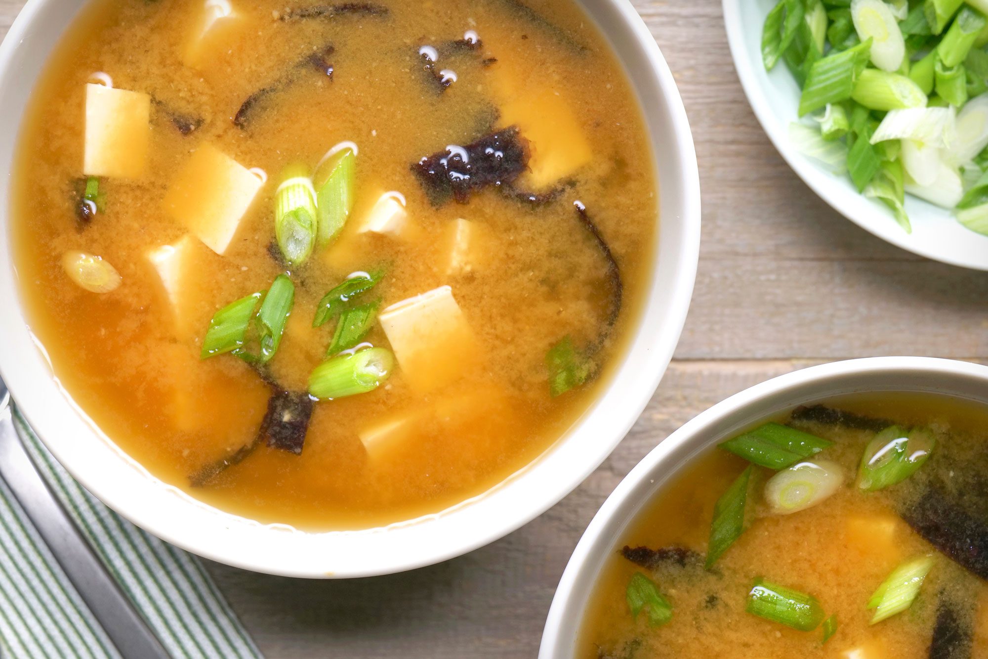 Overhead shot of Miso Soup; Ladle into bowls; garnish with green onion; spoon; napkin; wooden surface;