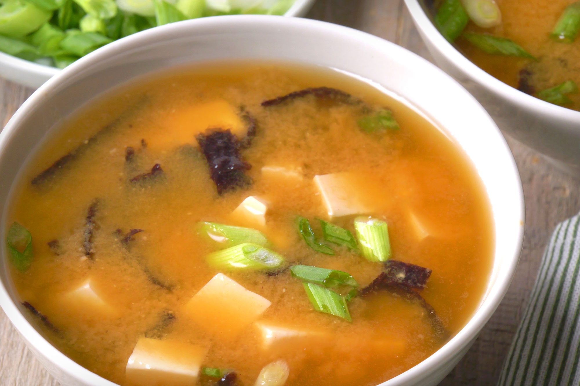 Close shot of Miso Soup; Ladle into bowls; garnish with green onion; spoon; napkin; wooden surface;