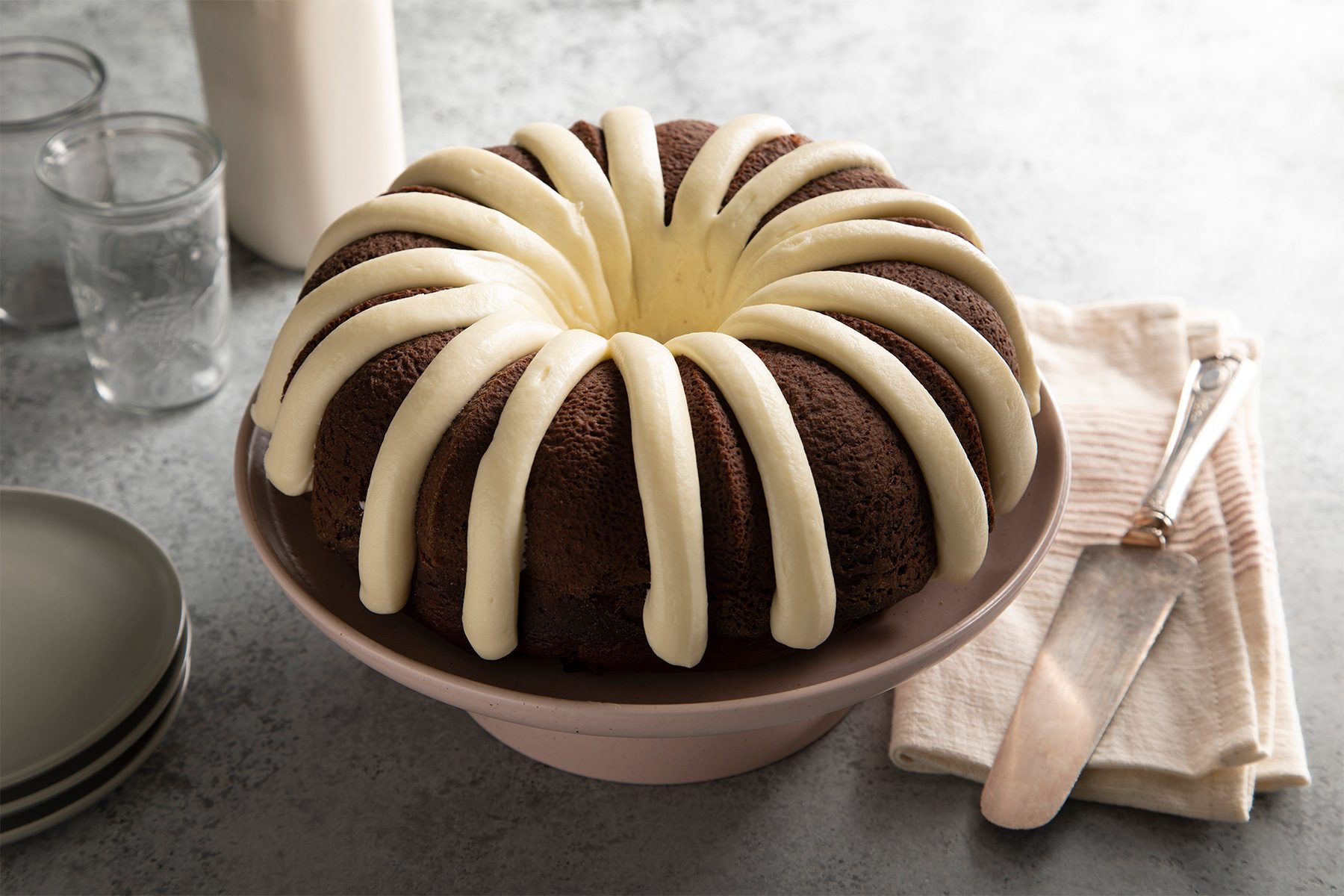 A chocolate bundt cake decorated with white icing drizzled in a radial pattern. It's placed on a cake stand beside a stack of plates, a knife, and napkins. Two glasses and a bottle are in the background on a gray surface.