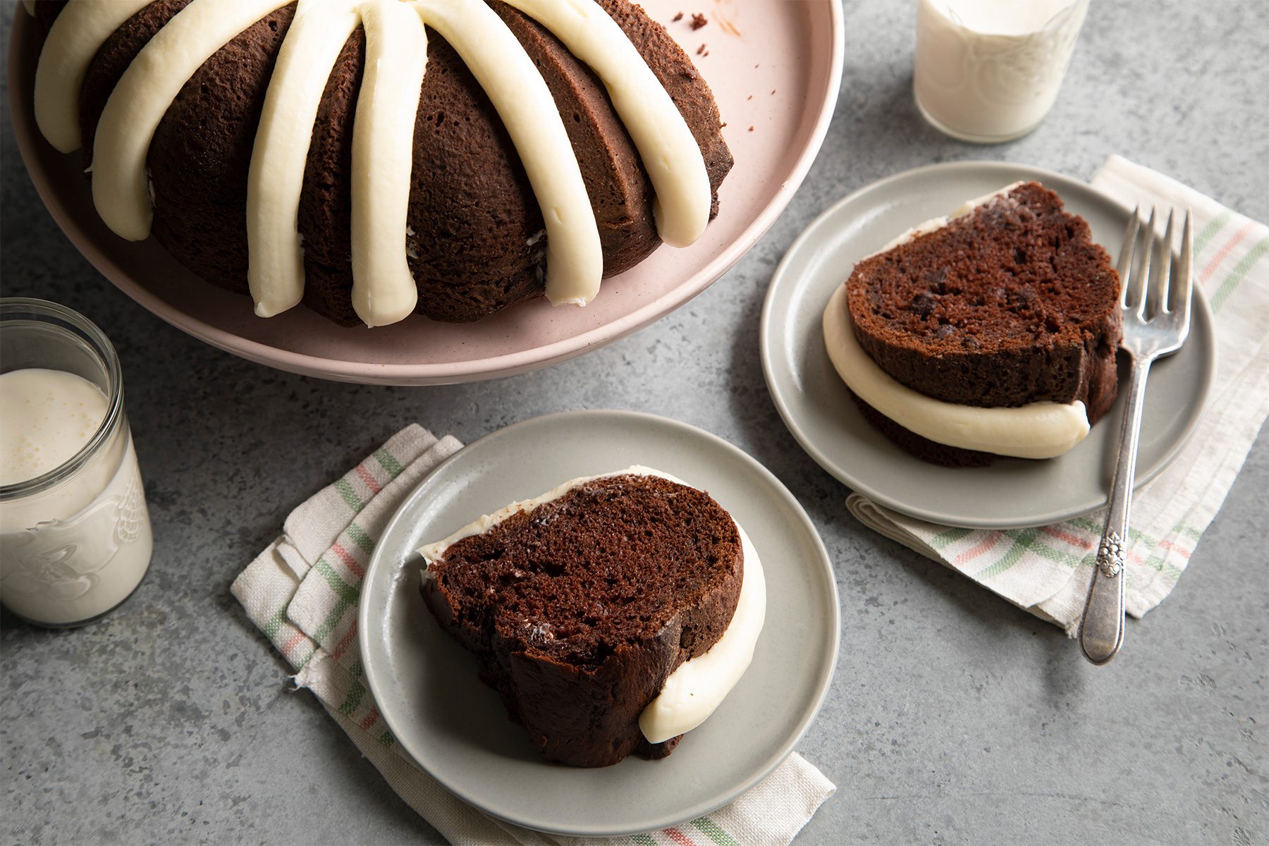 A chocolate bundt cake with cream cheese frosting on a pink plate. Two slices are served on gray plates with forks. A glass of milk and a jar of sugar are nearby on a gray surface with striped cloth napkins.