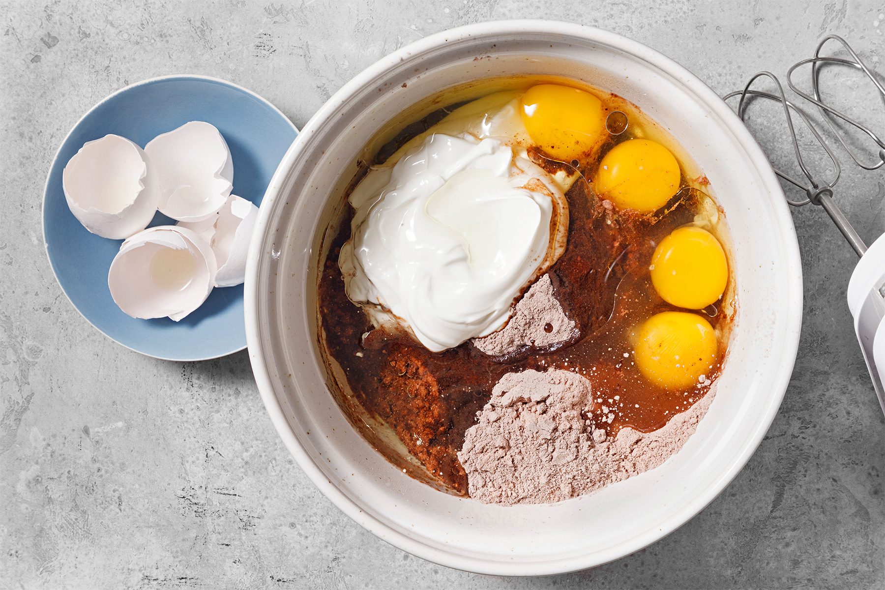 overhead shot of a white mixing bowl sits on a grey countertop; the bowl is filled with the ingredients for a cake batter or other baked good; the ingredients include a dollop of sour cream, four egg yolks, a brown liquid, and some cocoa powder; a blue plate sits to the left of the bowl; it holds three broken eggshells; a silver hand mixer sits on the right side of the bowl