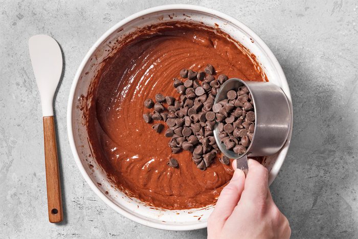 overhead shot of a white bowl sits on a grey countertop with a hand pouring chocolate chips into the bowl from a measuring cup; the bowl contains a dark chocolate batter; a spatula with a white blade and a brown handle sits to the left of the bowl; the hand is holding the measuring cup over the bowl and is pouring chocolate chips into the batter