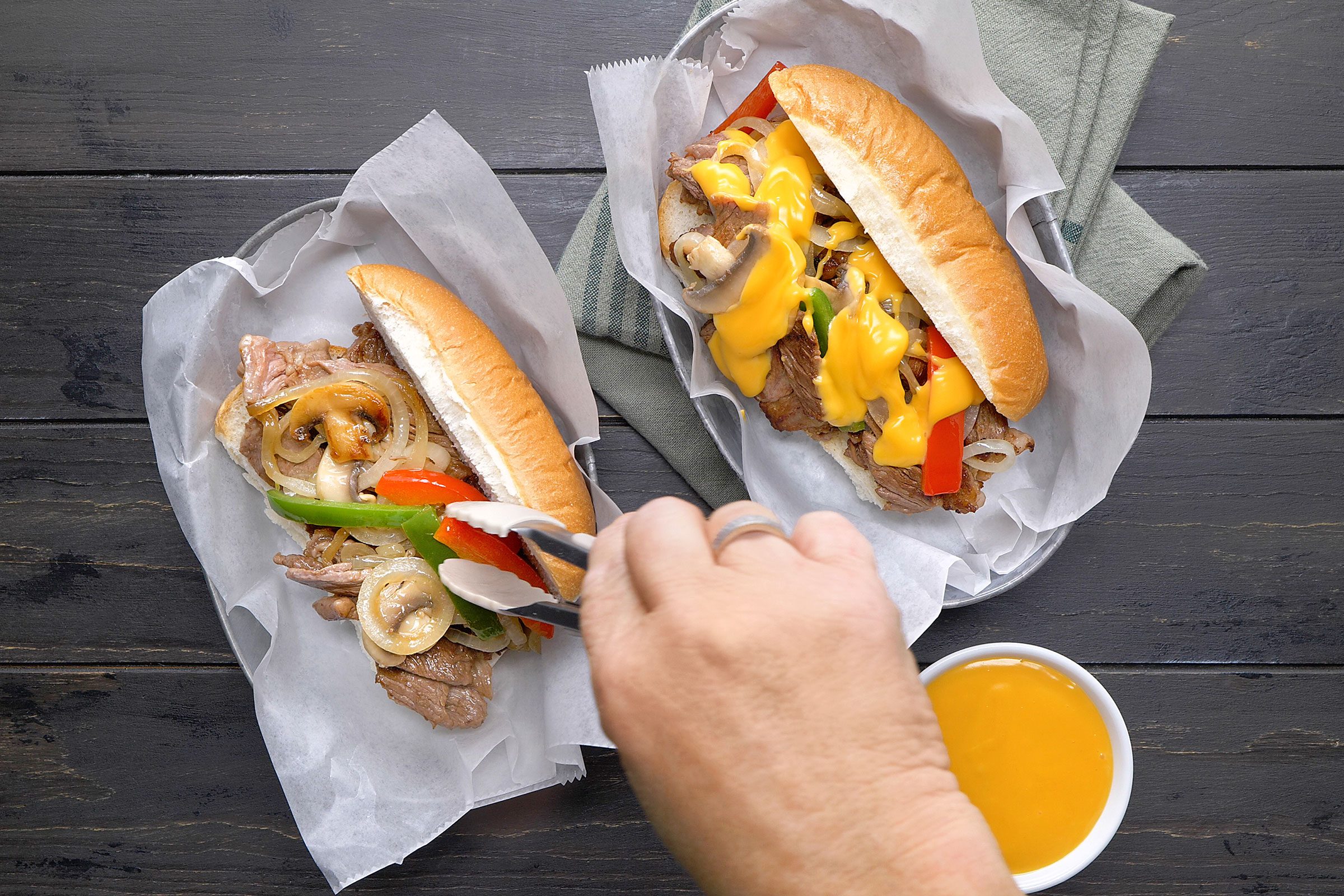 Assembling philly cheesesteak by adding ribeye steak in sliced roll topped with vegetables and cheese sauce